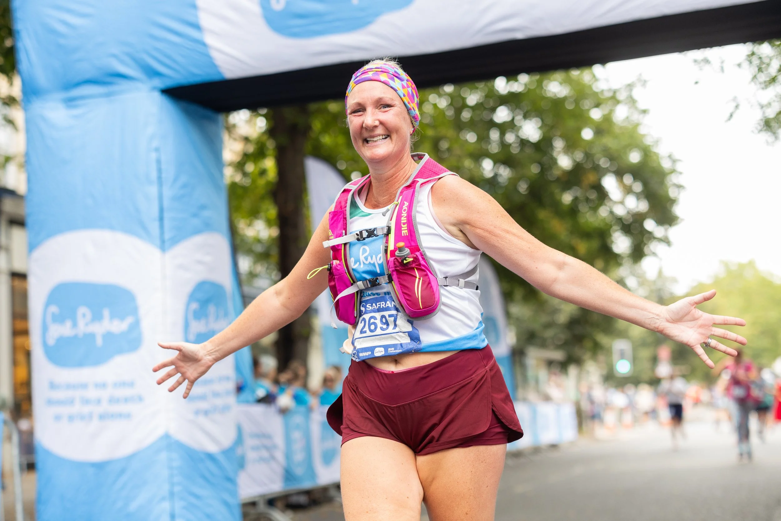 A smiling woman crossing the finish line at a race, wearing a colorful headband, a white sleeveless top, maroon shorts, and a pink hydration pack, with her arms outstretched.
