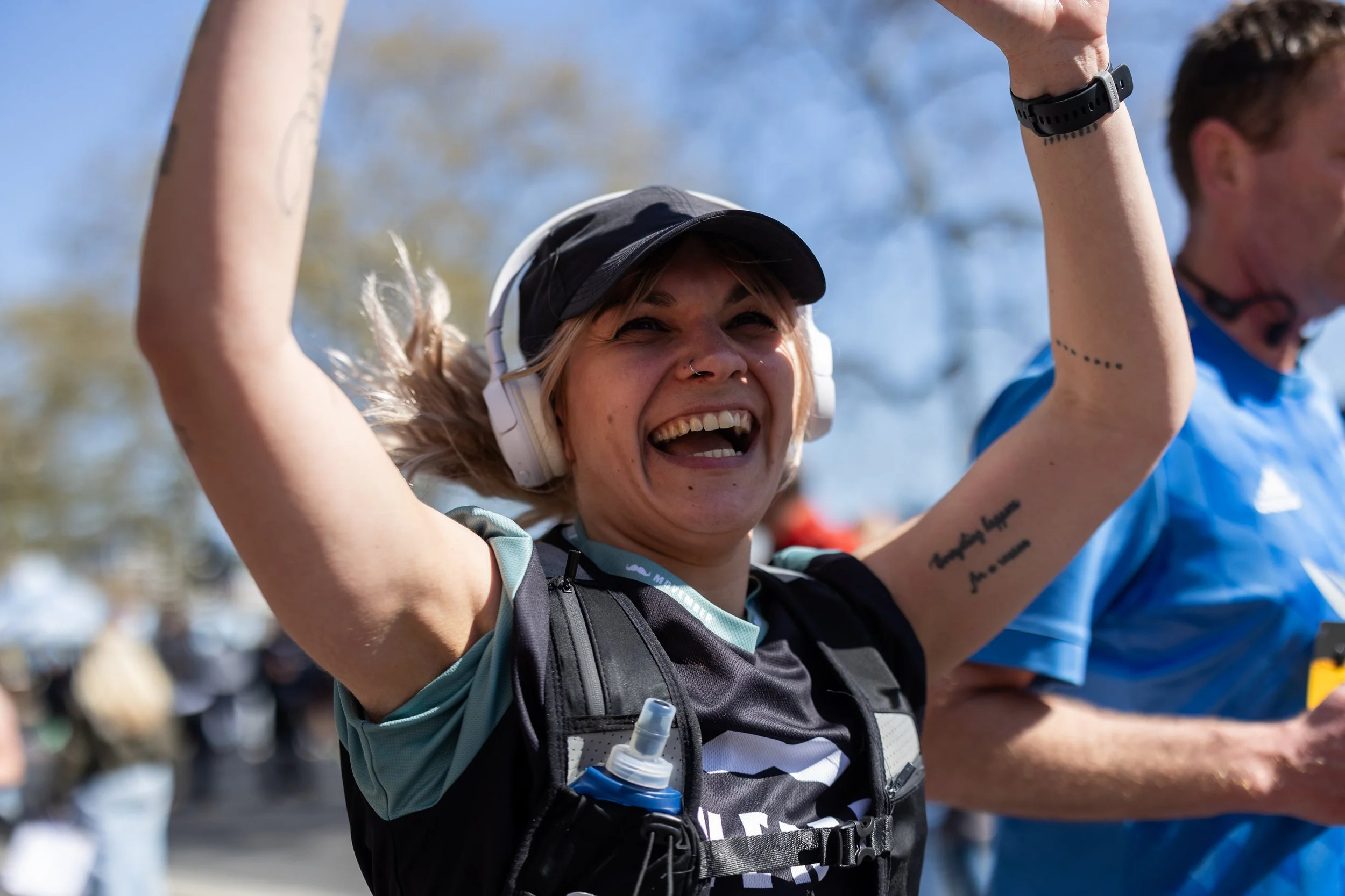 Woman smiling and raising her arms at an outdoor event, wearing a black cap, white headphones, and a running vest, with a person in a blue shirt beside her.