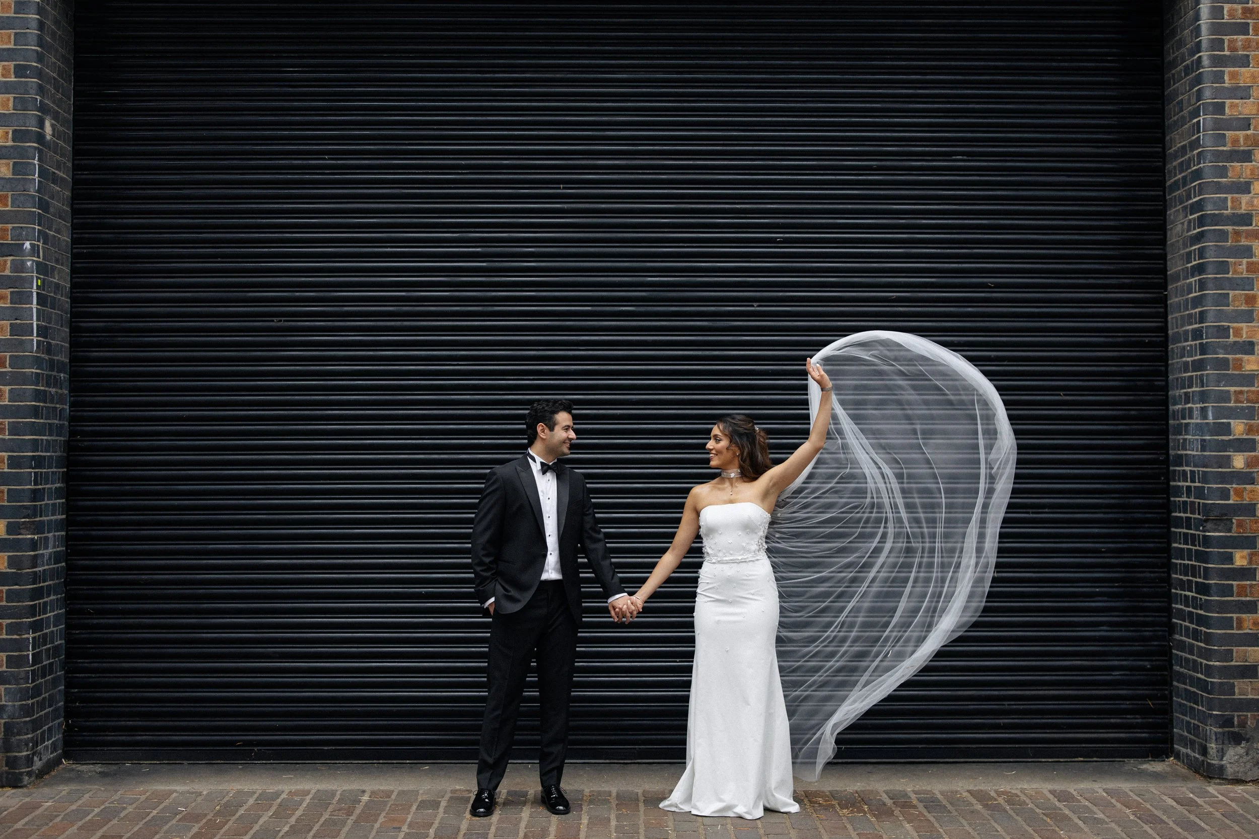 A bride waves her veil in the air artistically as she poses with her groom for wedding photos in Shoreditch