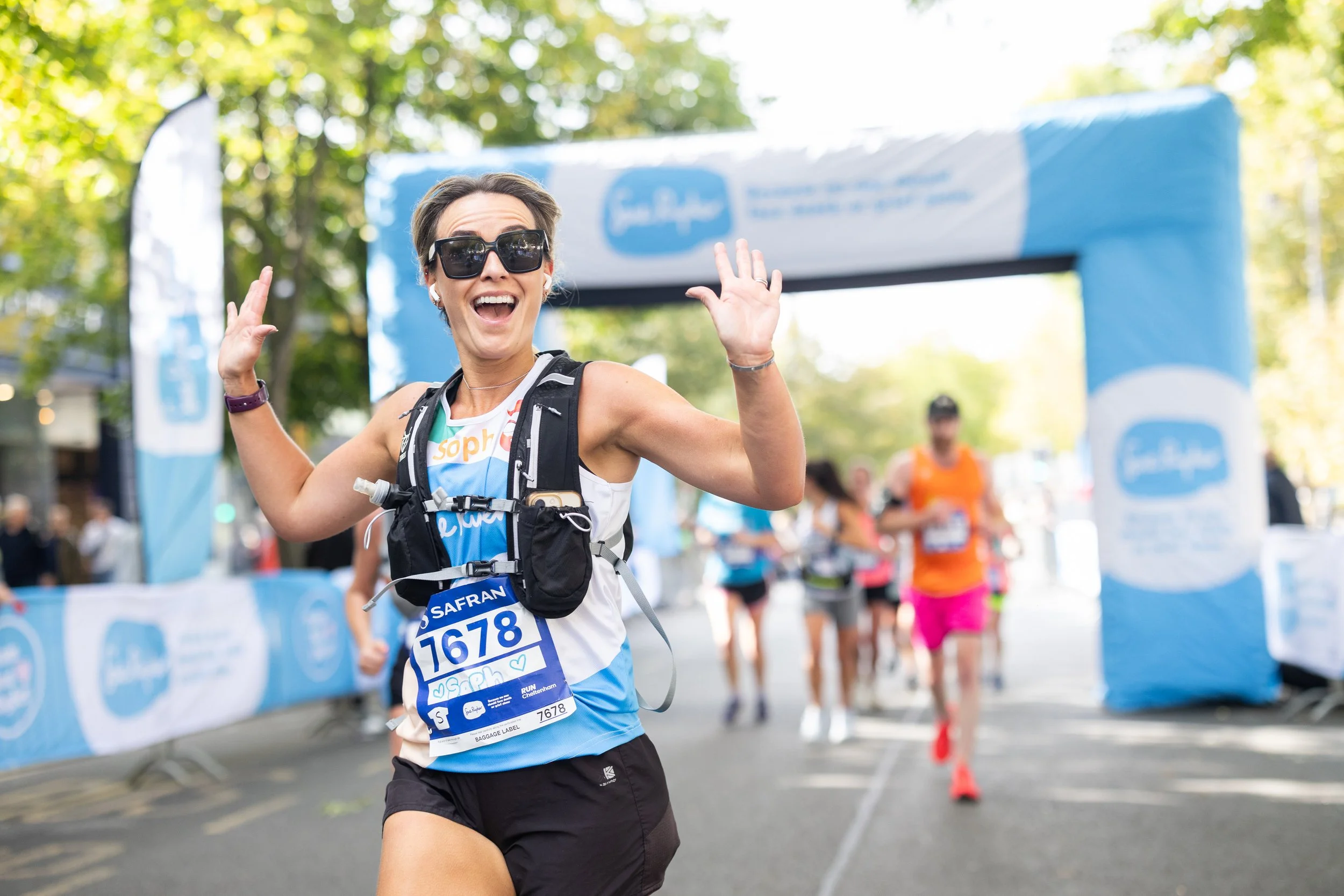 A female marathon runner smiling and waving as she crosses the finish line with other runners behind her, on a street lined with trees and blue race banners.