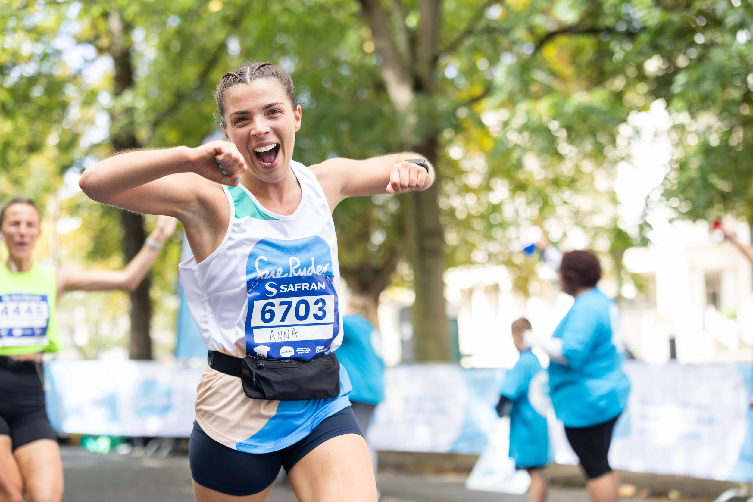 A smiling female marathon runner celebrating near the finish line, wearing a white and blue tank top with the number 6703 and the name Anna, with trees and other participants in the background.