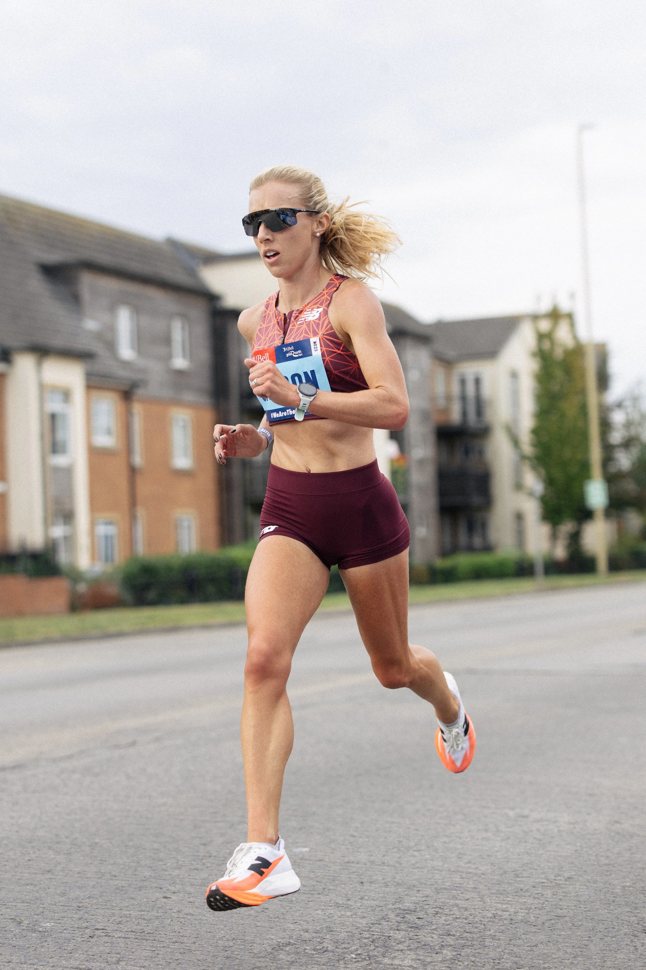 Female marathon runner in athletic gear and sunglasses running on street with residential buildings in background.