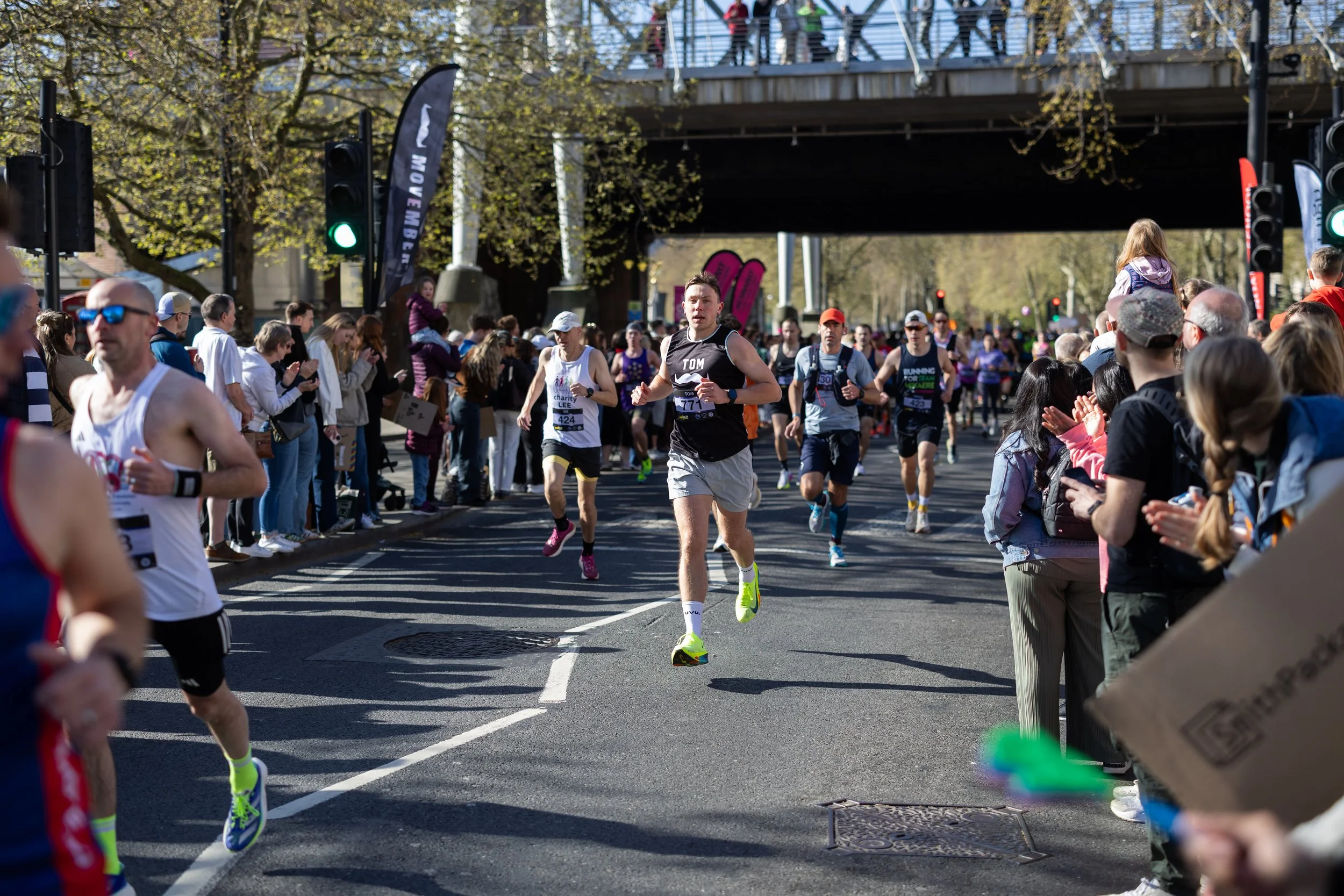 Marathon runners competing on city street with spectators on sides, under a bridge on a sunny day.