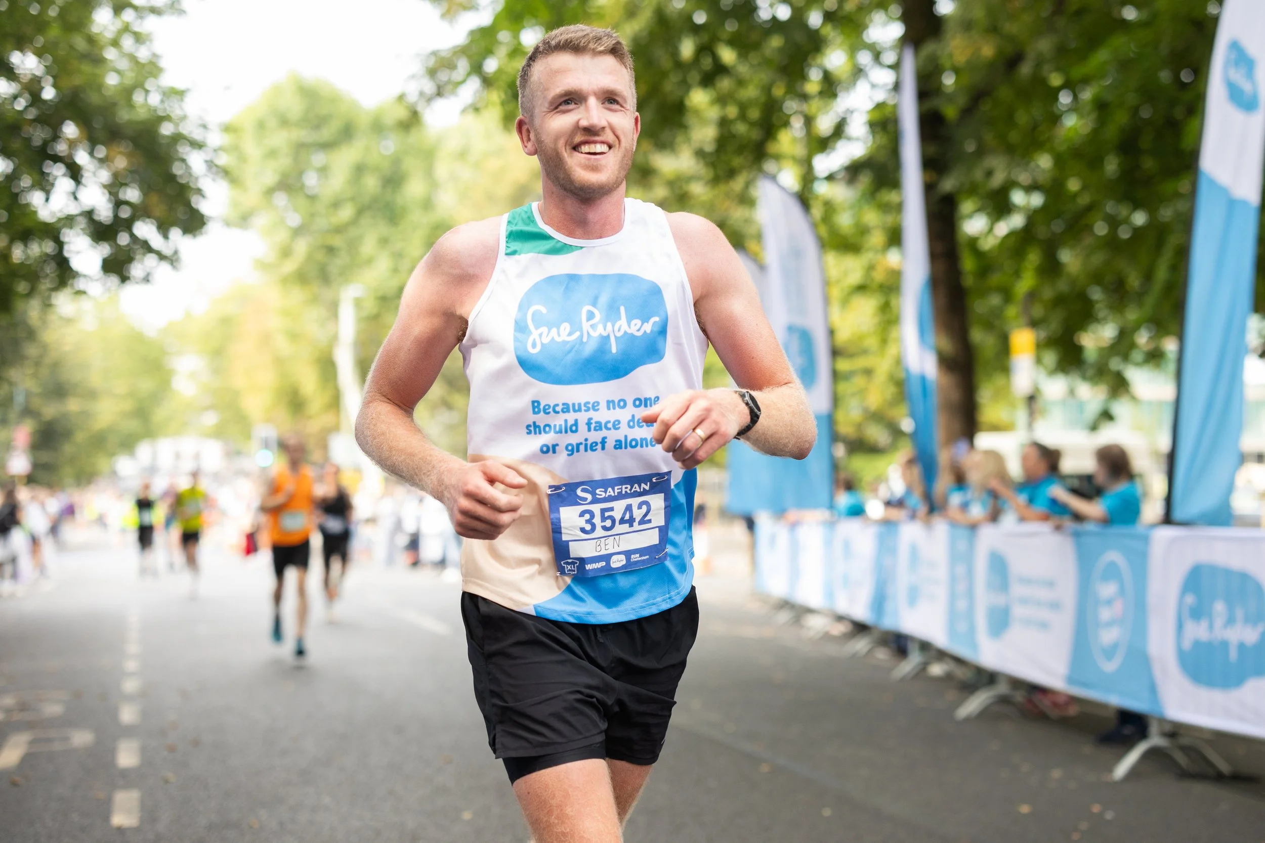 A man running in a marathon, smiling, wearing a race bib with the number 3542 and the name Ben, with trees and race banners in the background.