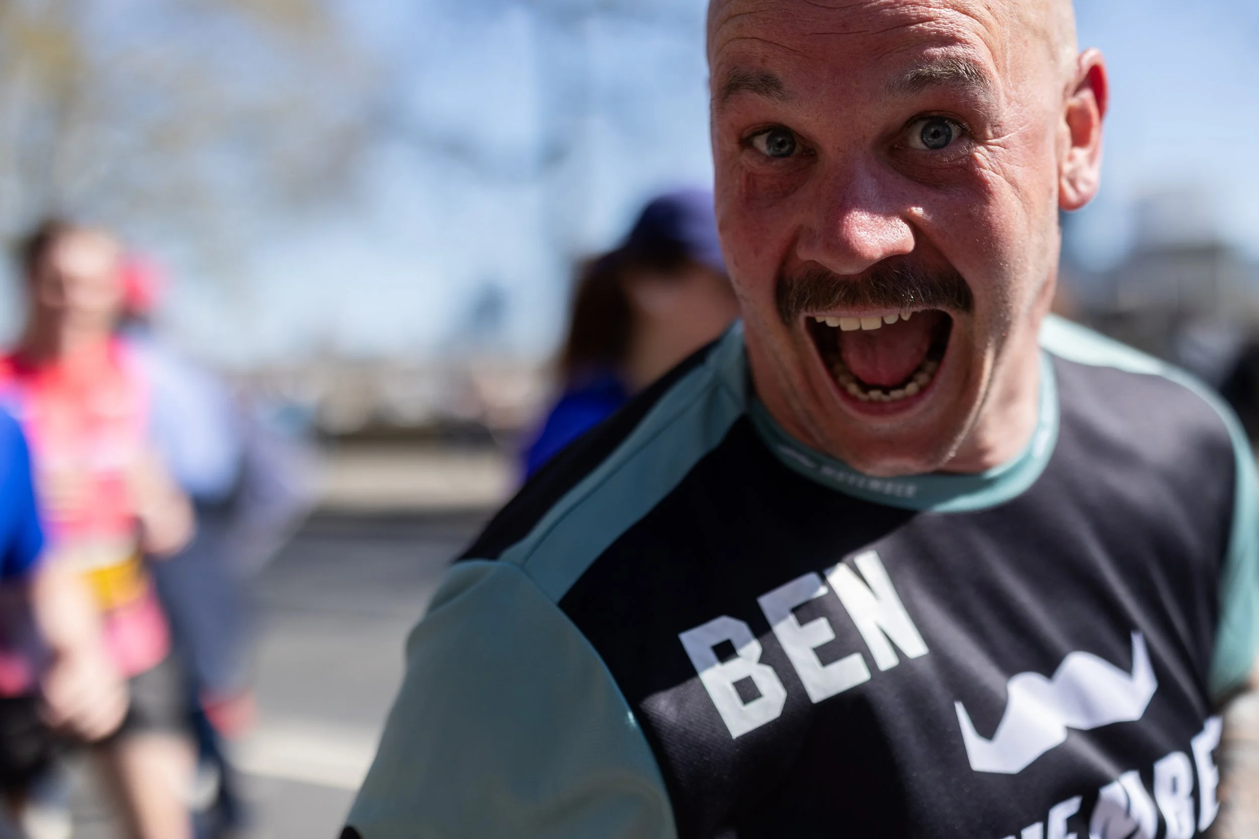 Man with a bald head and mustache shouting with an open mouth, wearing a black and light blue shirt, during a race or marathon.