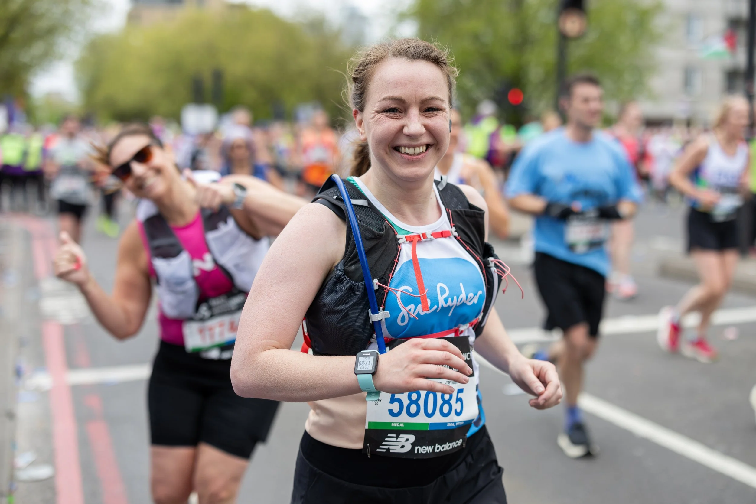 A smiling woman running in a marathon, wearing a race bib and athletic gear, surrounded by other runners on a city street.