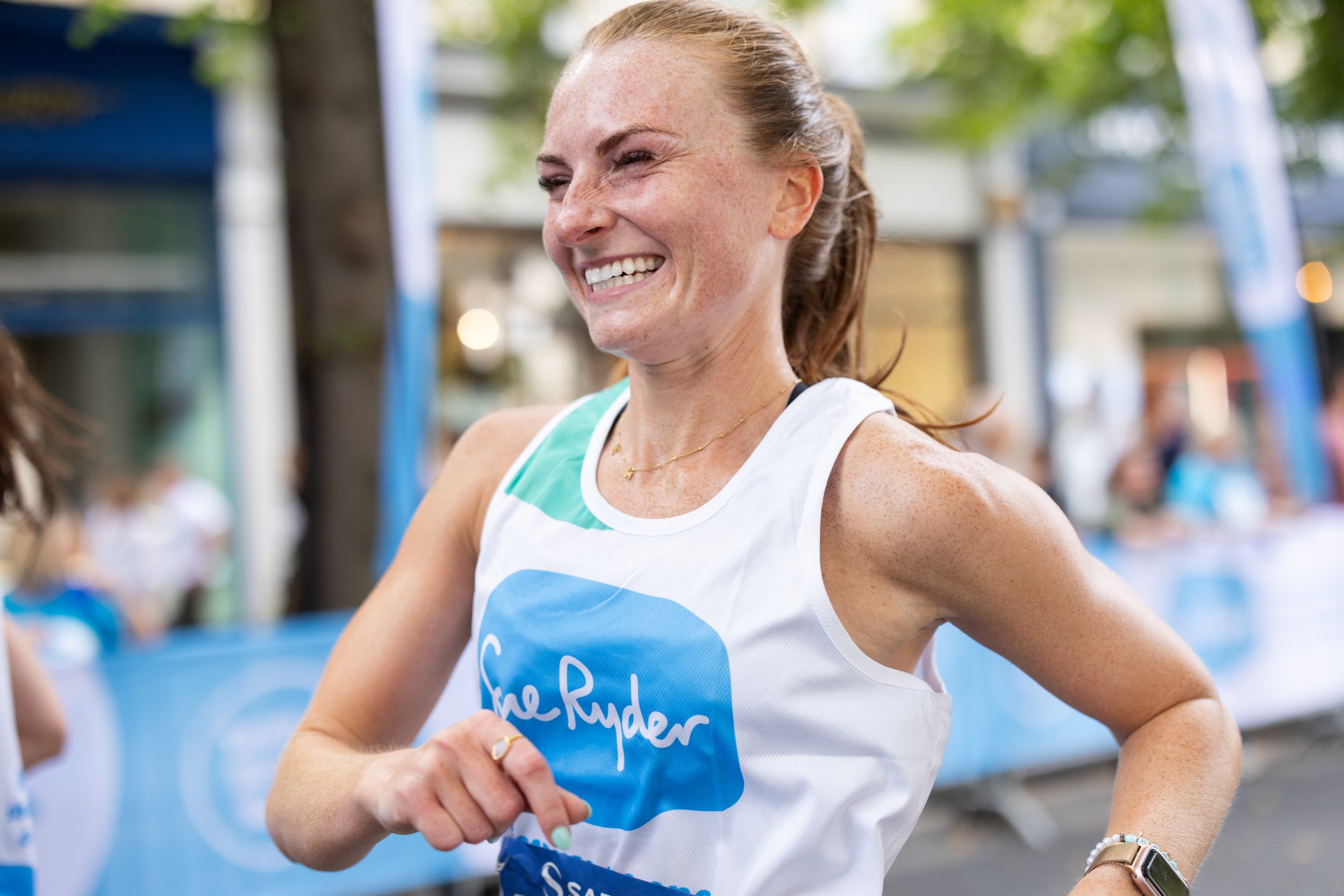 A woman running in a race, smiling, wearing a white tank top with a blue logo and a gold necklace, with trees and race banners in the background.