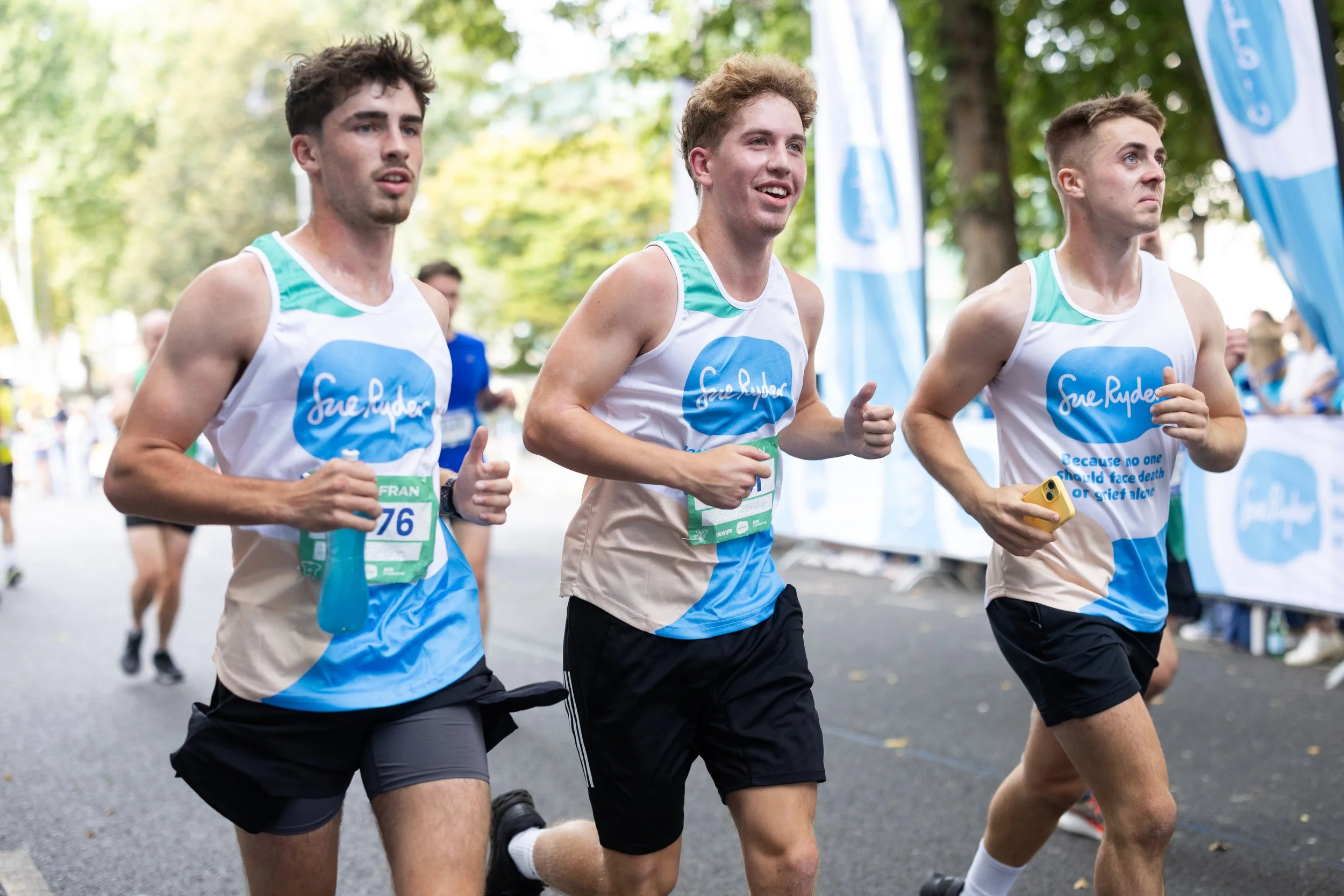 Three male runners participating in a race, wearing matching shirts with a logo and quote, running on a street lined with trees and blue banners.
