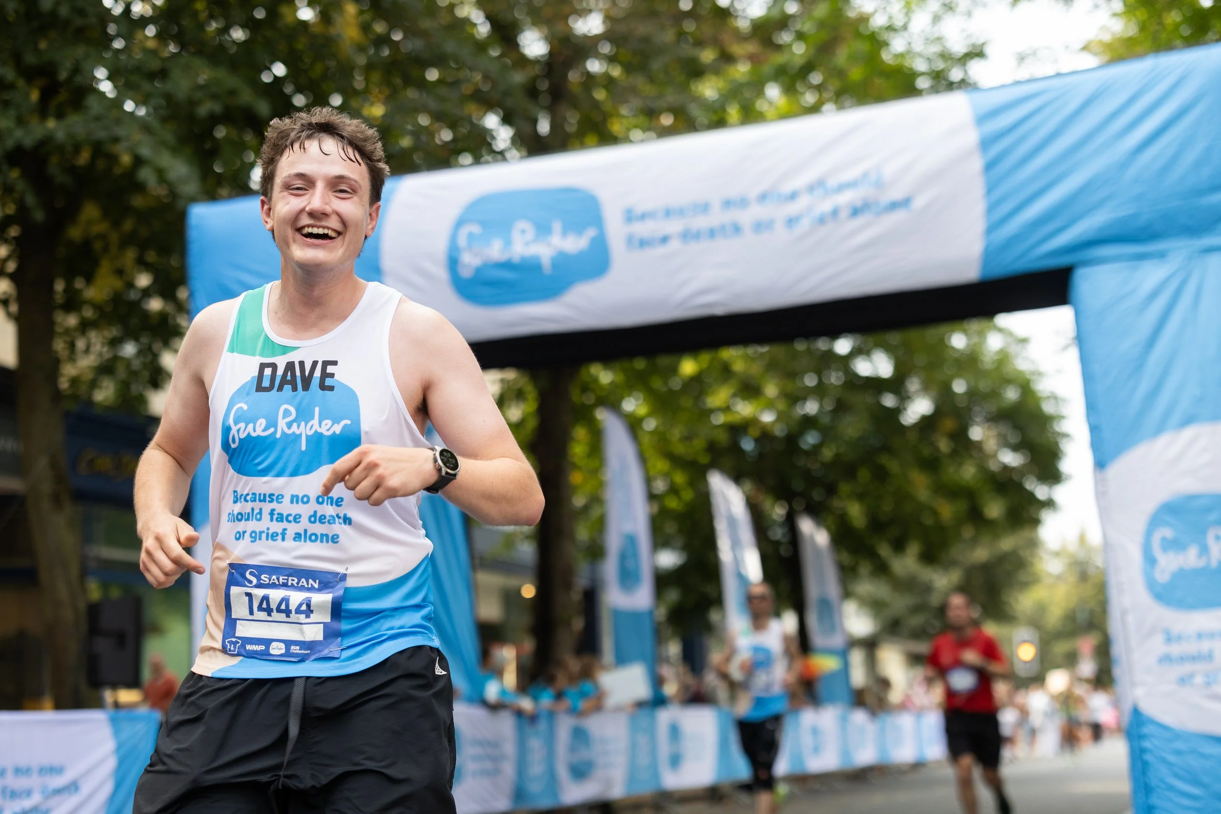 Male runner smiling and crossing the finish line at a race, wearing a bib number 1444 and a shirt with the name 'Sue Ryder' and the phrase 'Because no one should face death or grief alone'.