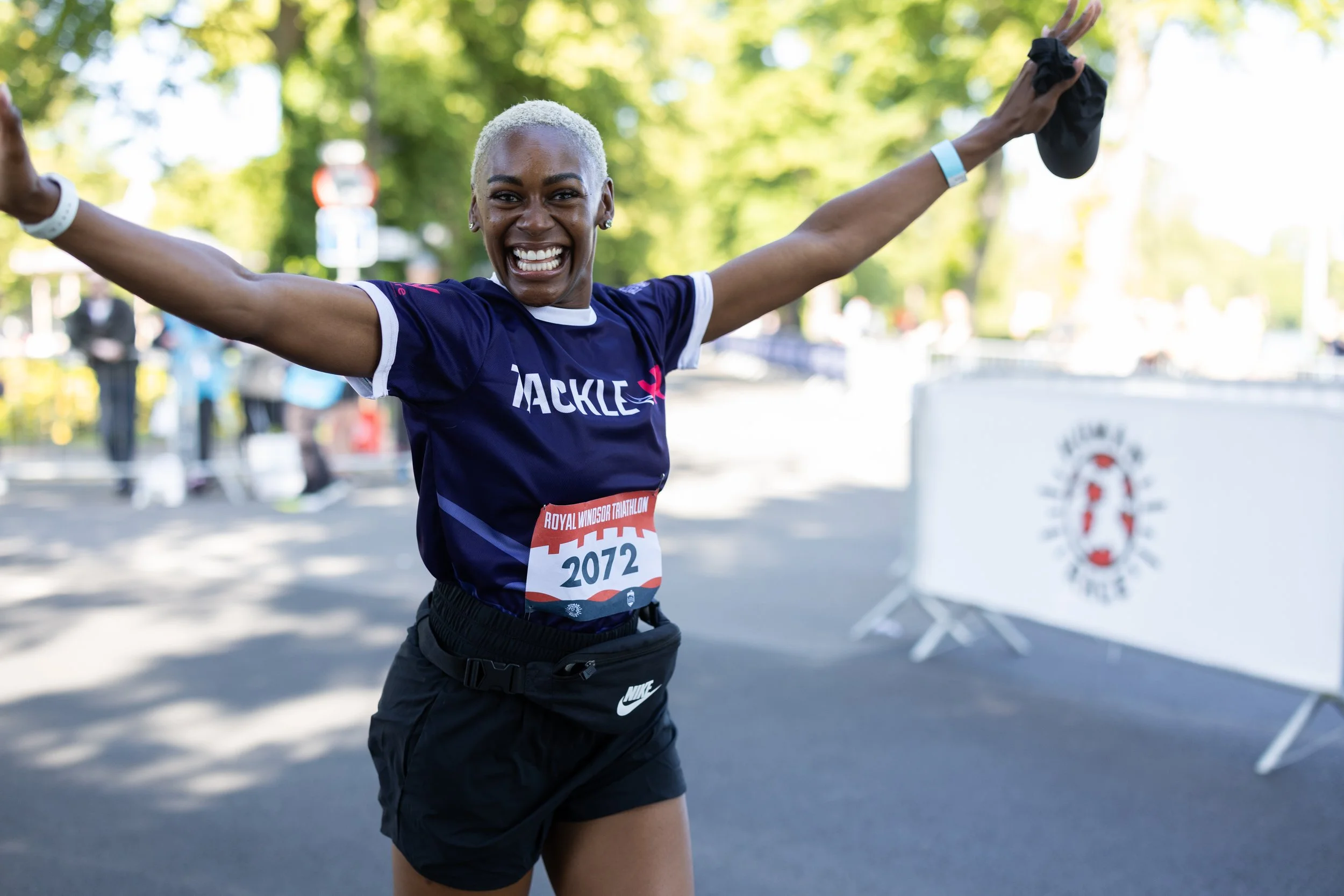 A smiling woman with short blonde hair celebrating at the finish line of a race, wearing a navy blue running shirt, black shorts, and a bib with the number 2072.