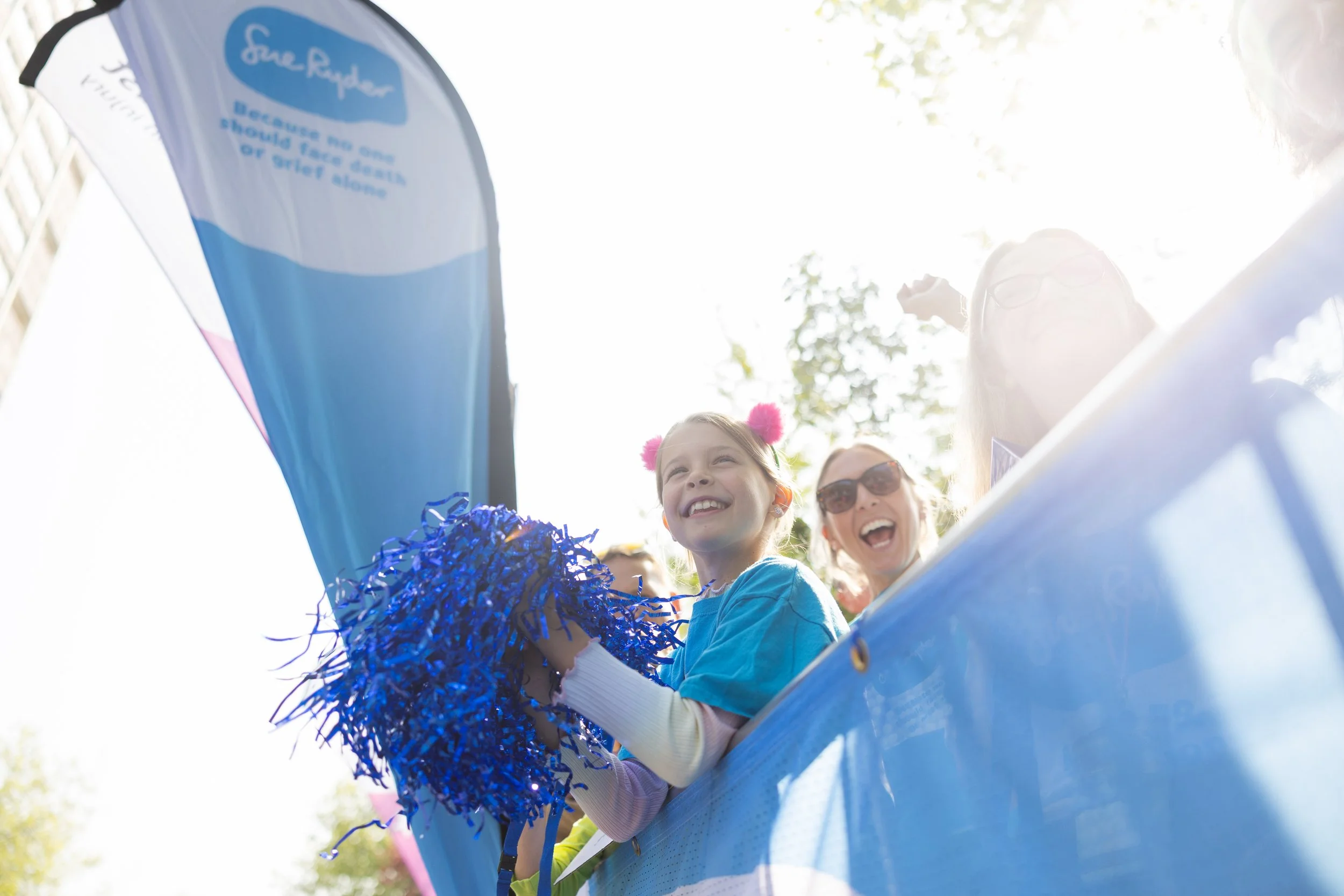 People celebrating at a parade or event, with a smiling girl holding blue pom-poms, and others around her laughing and enjoying the moment, with bright sunlight and flags in the background.