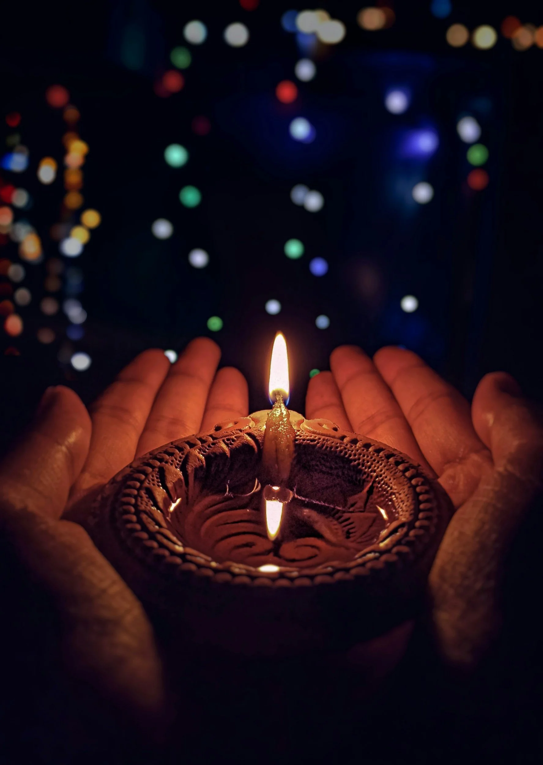 Person holding a small clay diya lamp with lit wick, surrounded by outstretched hands, with colorful blurred holiday lights in the background.