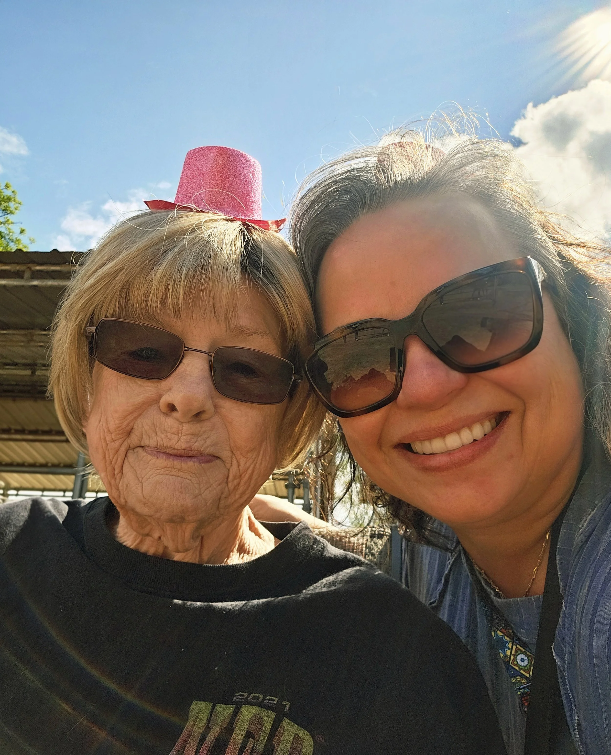 A close-up selfie of two women with sunglasses outdoors in bright sunlight. One woman appears to be elderly with light-colored hair, and the other woman is middle-aged with dark hair. The sky is clear with some clouds, and there is a rainbow-like lens flare effect in the photo.