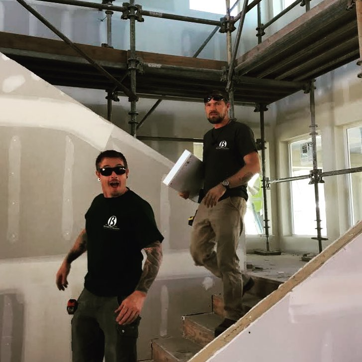 Two men working on a construction site inside a house with scaffolding and drywall. One man is standing on a staircase holding a notebook, while the other man is standing on the stairs wearing sunglasses and smiling.