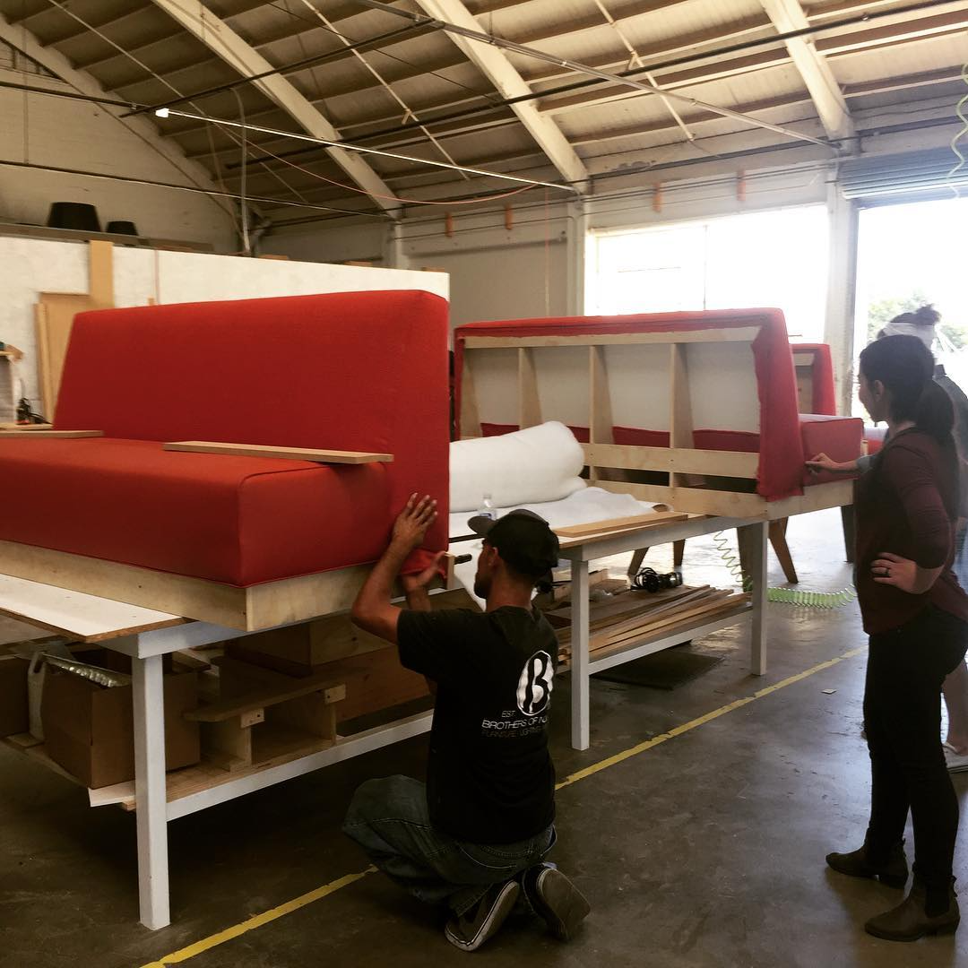People assembling red upholstered furniture in a workshop, with wooden frames and tools around.