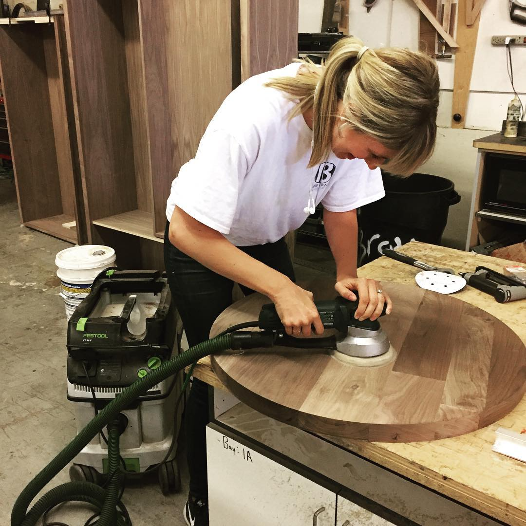 A woman using a handheld power sander on a large round wooden table in a woodworking workshop.