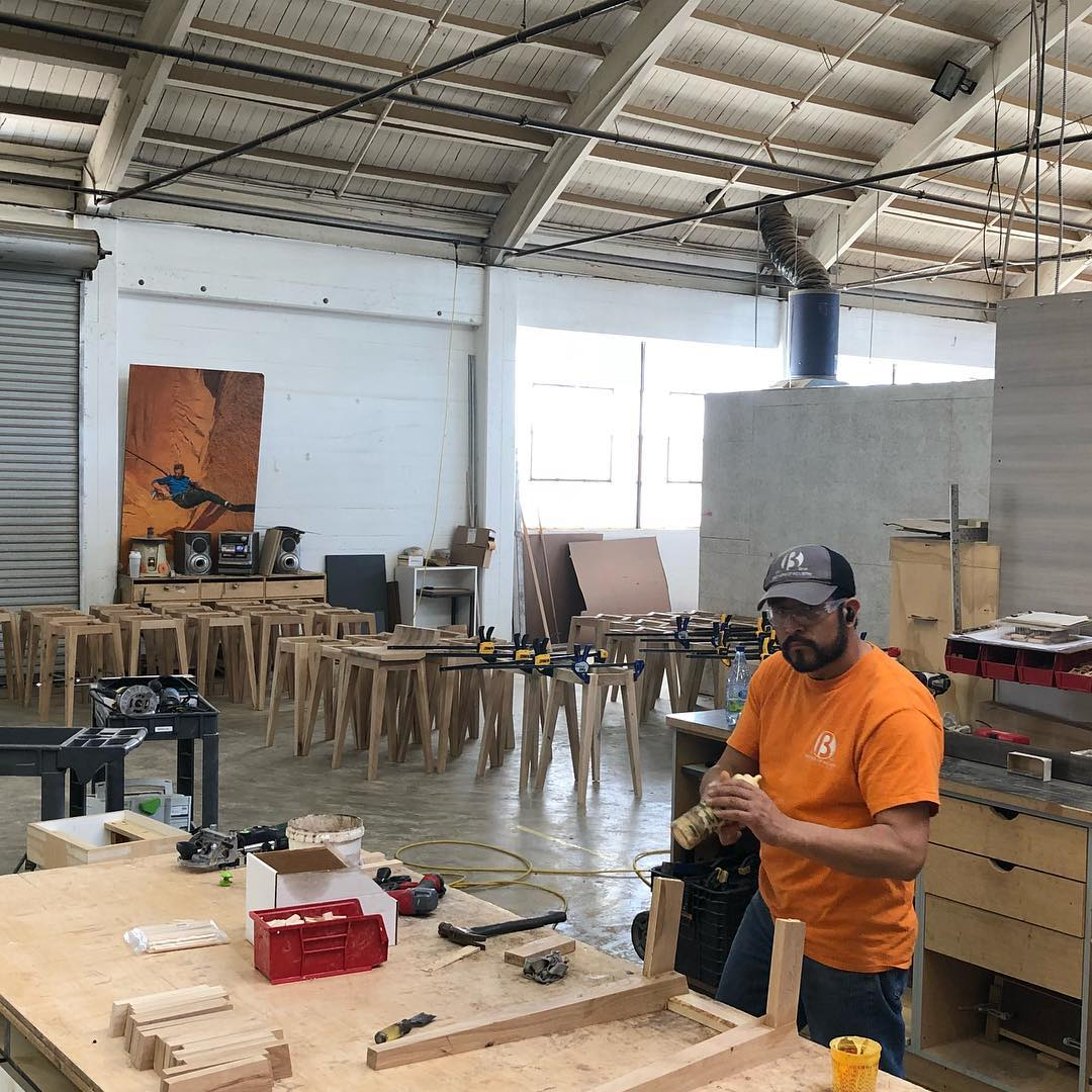 A man in an orange T-shirt working in a woodworking shop with wood pieces, clamps, and tools on a workbench, and multiple wooden stools lined up in the background.