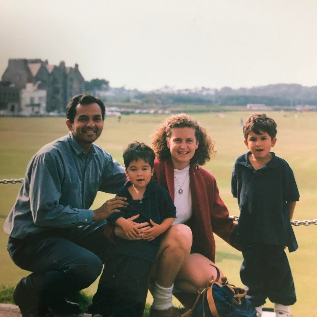 A family of four, two adults and two children, posing outdoors on a grassy field with large houses in the background during daytime.