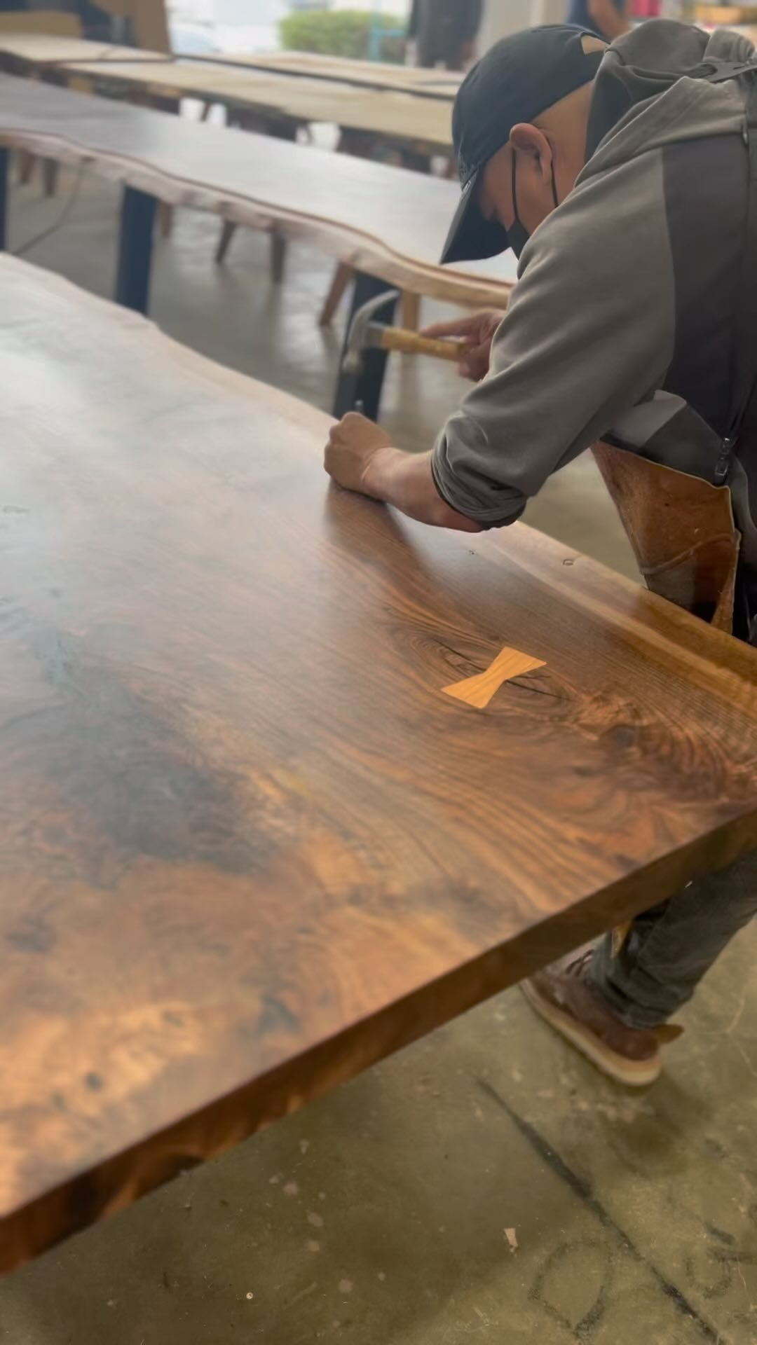 A person wearing a cap, glasses, a face mask, and a gray apron is working on a large wooden table in a workshop or carpentry shop, sanding or finishing the surface.