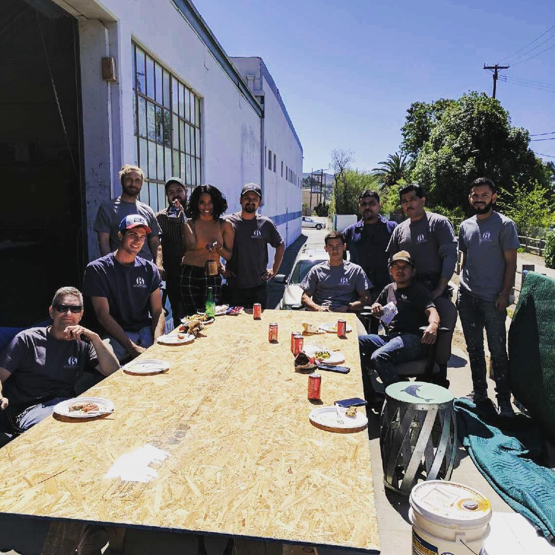 Group of twelve people gathered around an outdoor table with food and drinks, posing for a photo on a sunny day outside a building.