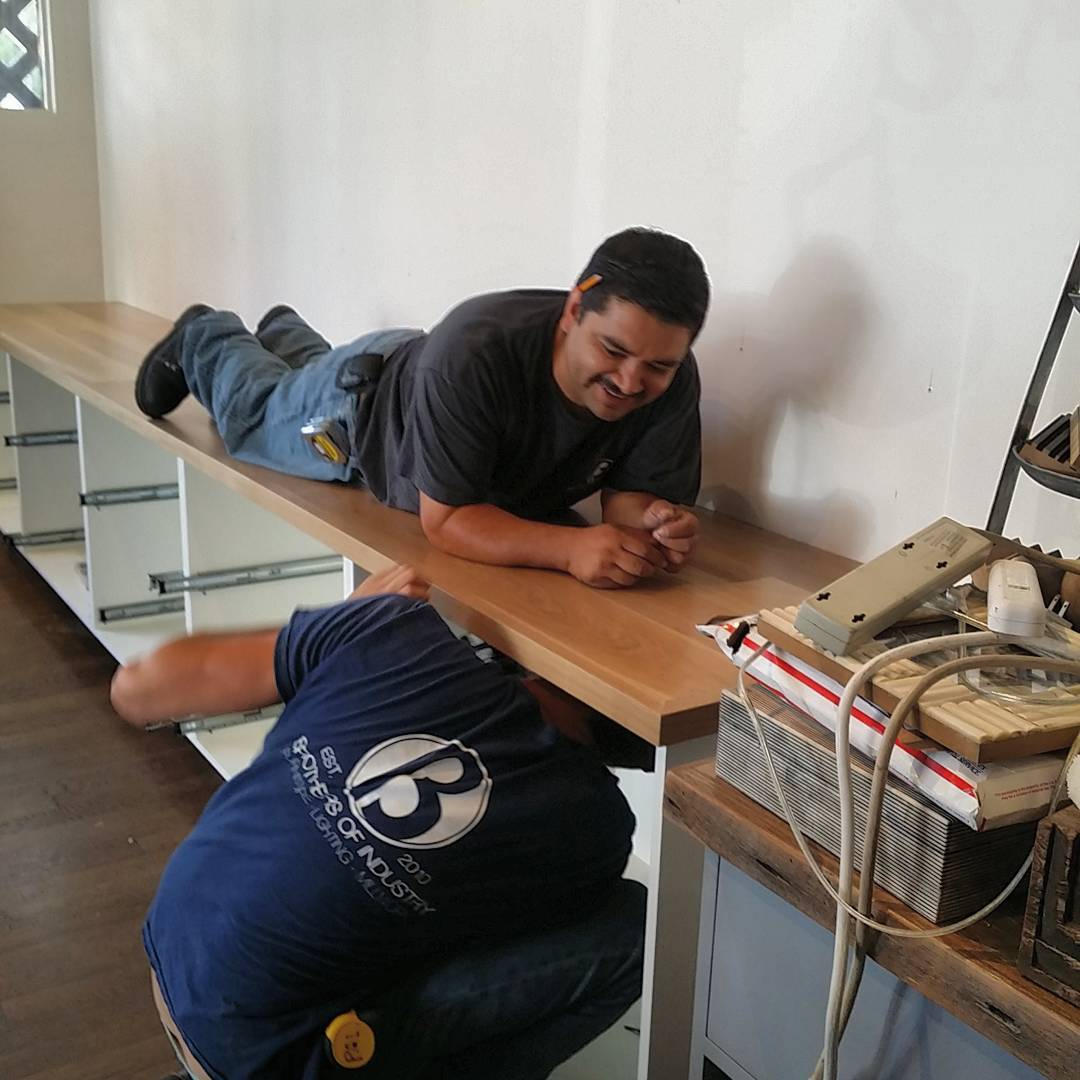 A man lying on a wooden counter, smiling, while another person crouches underneath, working on a cabinet or drawer.