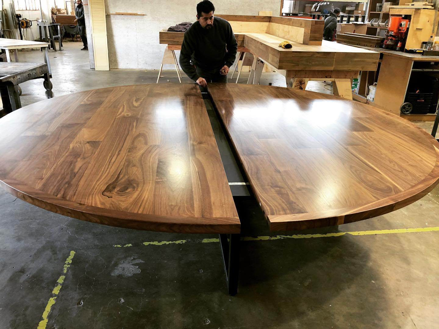 A large, handmade round wooden table in a workshop, with a person working on it and other workers in the background.