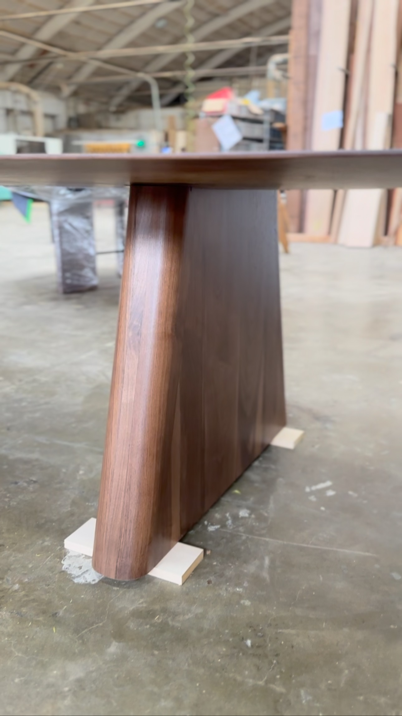 Close-up of a custom luxury sculpted walnut wooden table leg with a trapezoidal shape, showing the smooth dark brown wood grain walnut, in a woodworking shop or workshop with concrete floor and various woodworking materials in the background.