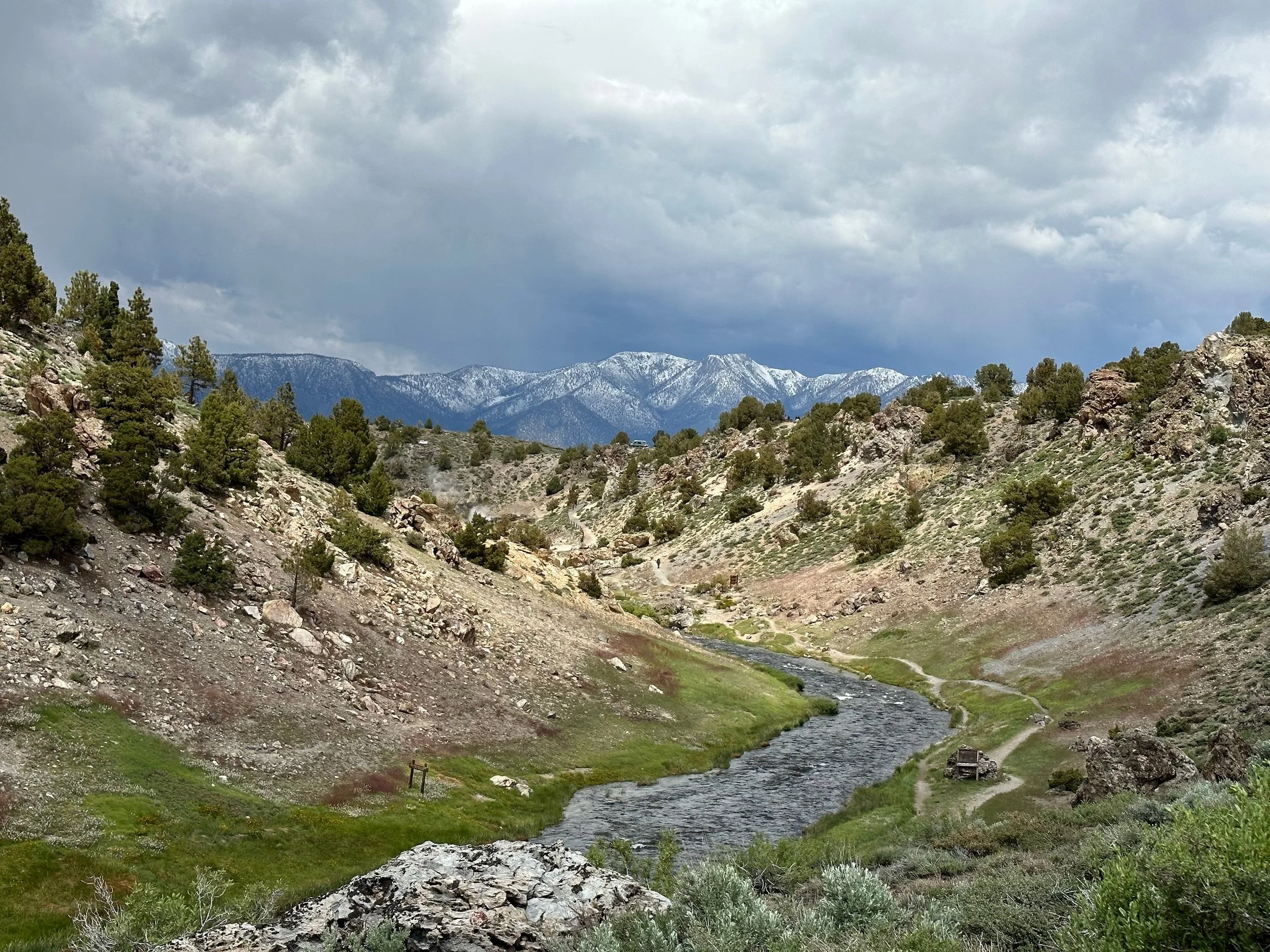 A scenic mountain landscape with a river flowing through a green valley, surrounded by rocky hills and sparse trees, with snow-capped mountains in the distance under a cloudy sky.