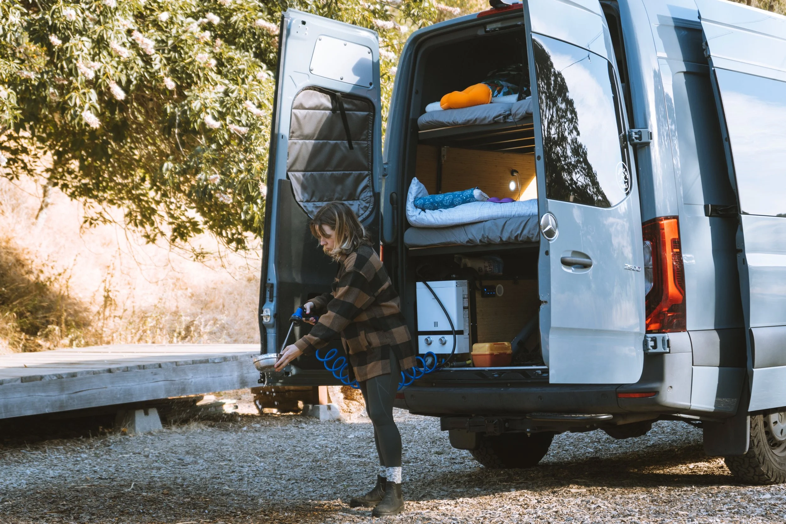 A young woman filling a camping stove outside a camper van with a bed visible inside on a gravel surface during daytime, with trees and sunlight in the background.