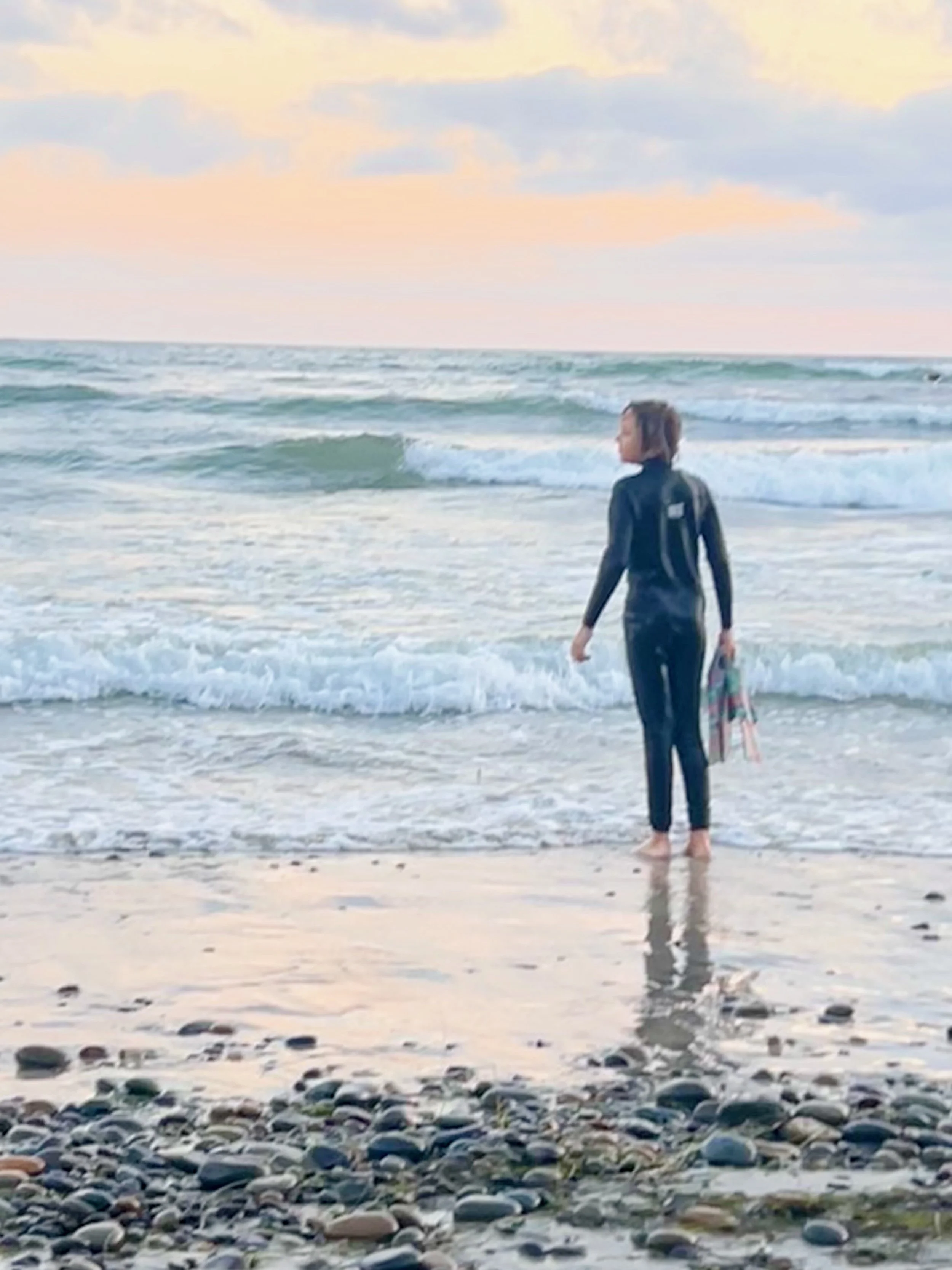 A person in a wetsuit standing at the edge of the ocean with waves and a pastel-colored sky in the background.