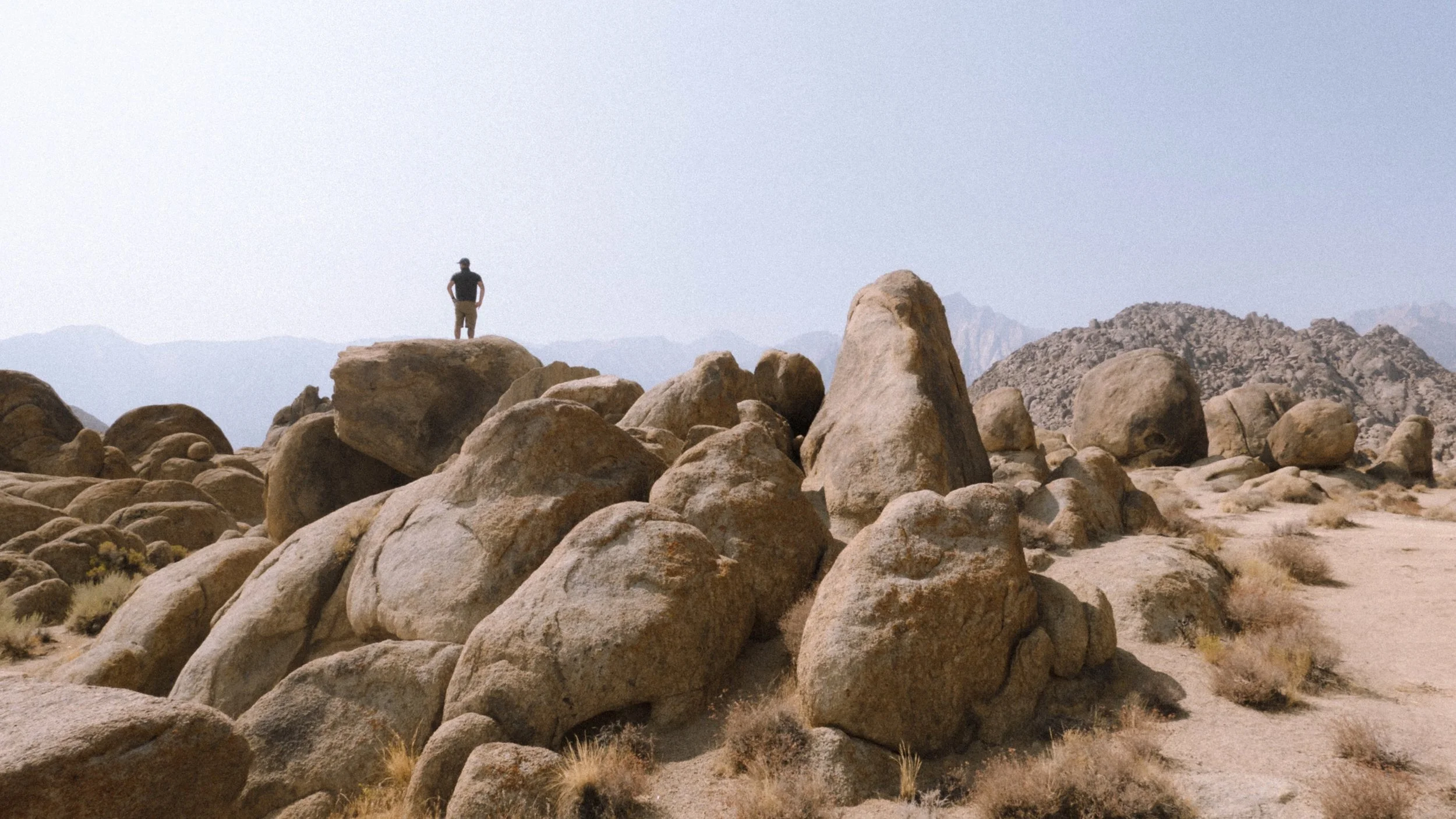 A man standing on a large rock in a desert landscape with scattered boulders and mountains in the distance.