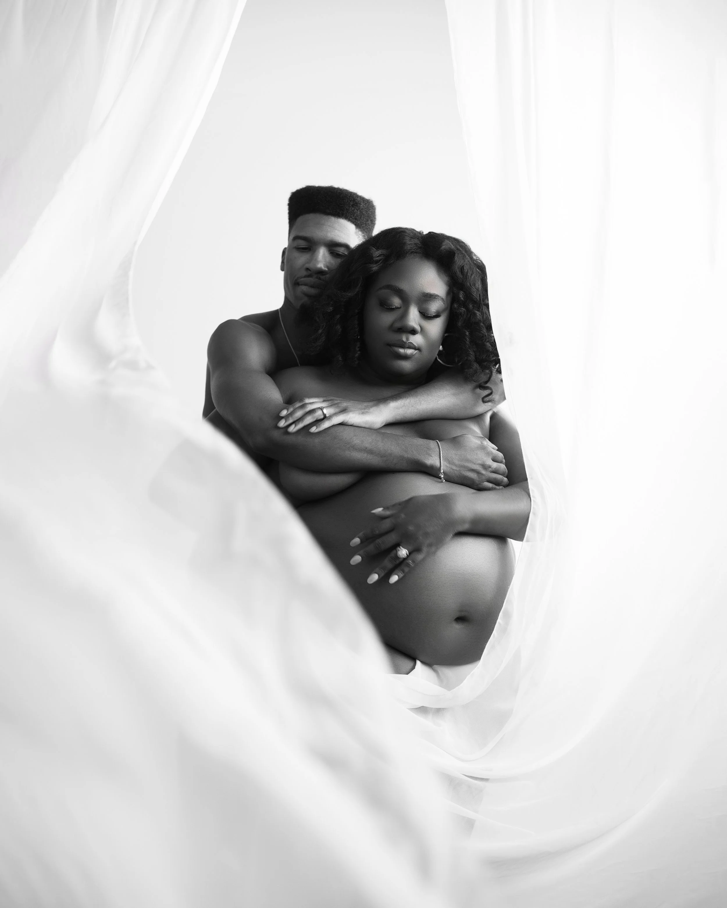 Black and white portrait of a pregnant woman with her partner embracing her from behind, both with eyes closed, surrounded by sheer white fabric.