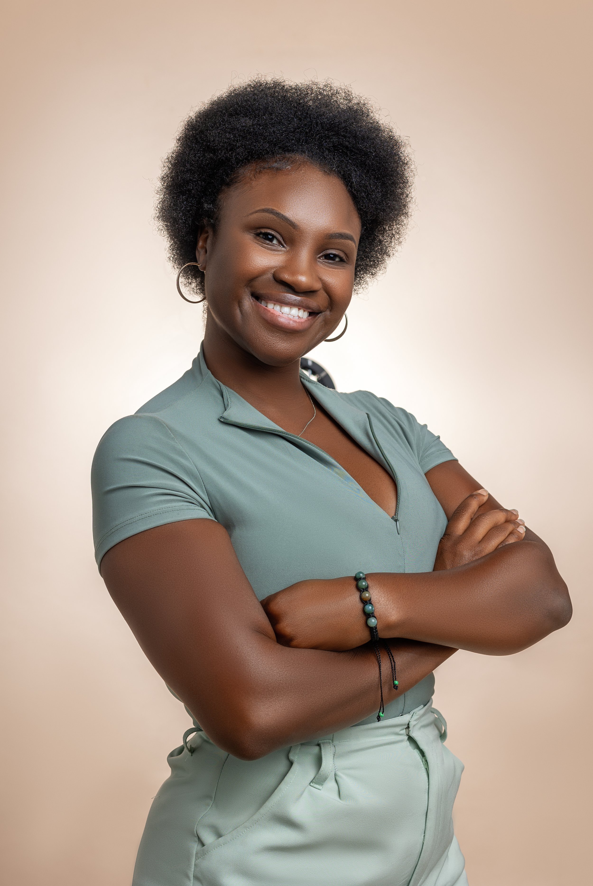 Stylish woman with curly hair smiling, wearing a fitted mintgreen t-shirt, hoop earrings, beaded bracelet, and posing with arms crossed.
