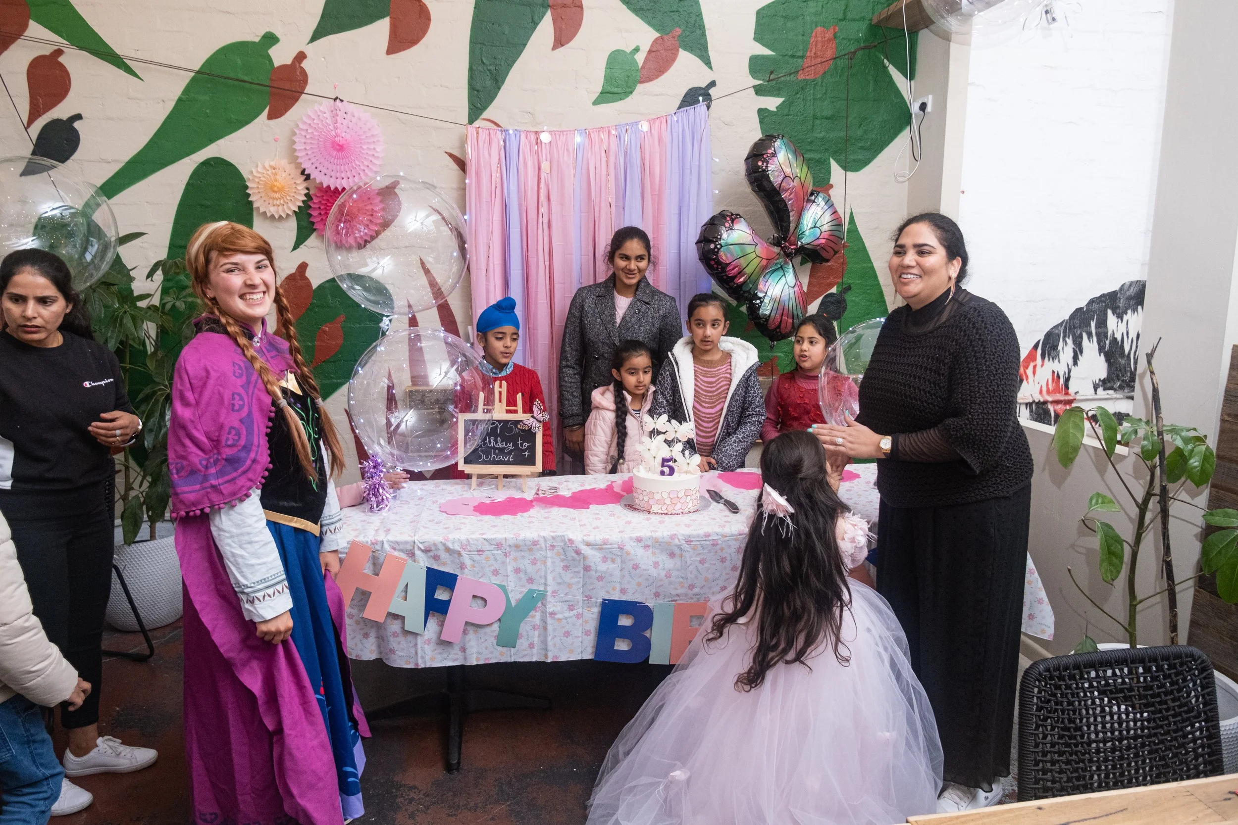 Children and adults gathered at a birthday celebration with pink, purple, and black decorations, including a birthday cake with the number 5, balloons, and a colorful butterfly-shaped balloon.