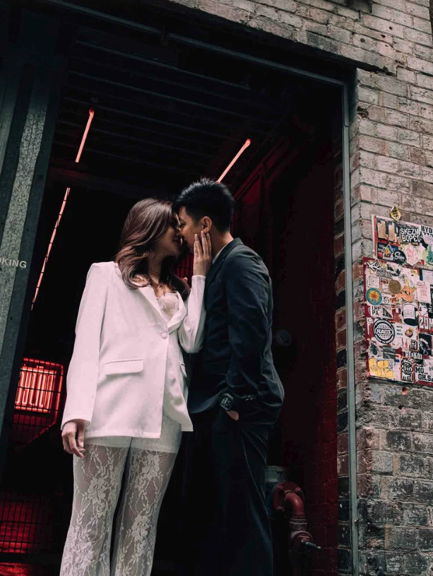 A couple leaning in for a kiss in an urban alleyway with brick walls and posters, illuminated by red and pink neon lighting.