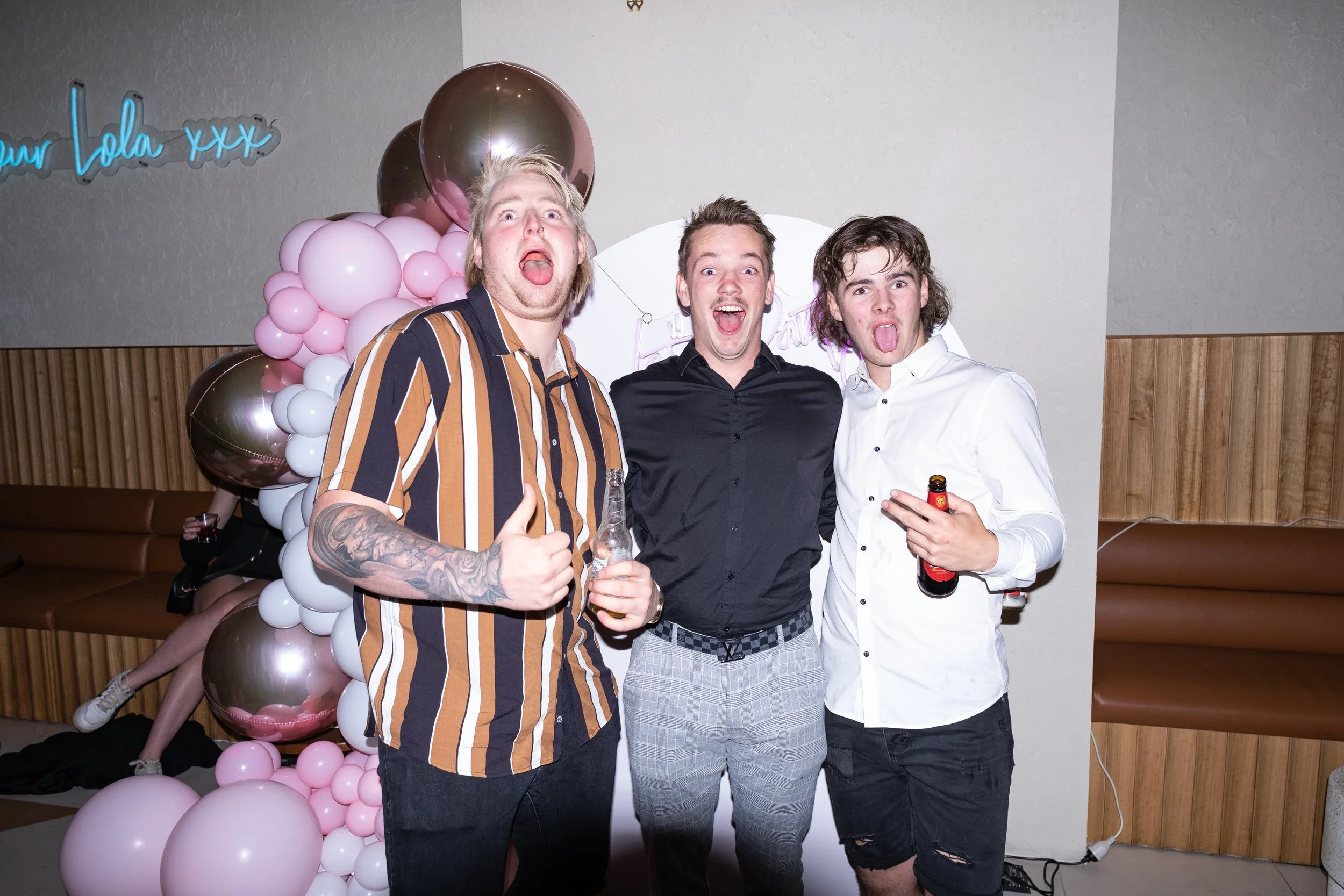 Three young men at a party posing with playful expressions, sticking out their tongues, in front of a pink and gold balloon decoration and a neon sign that reads 'your Lola XXX'.