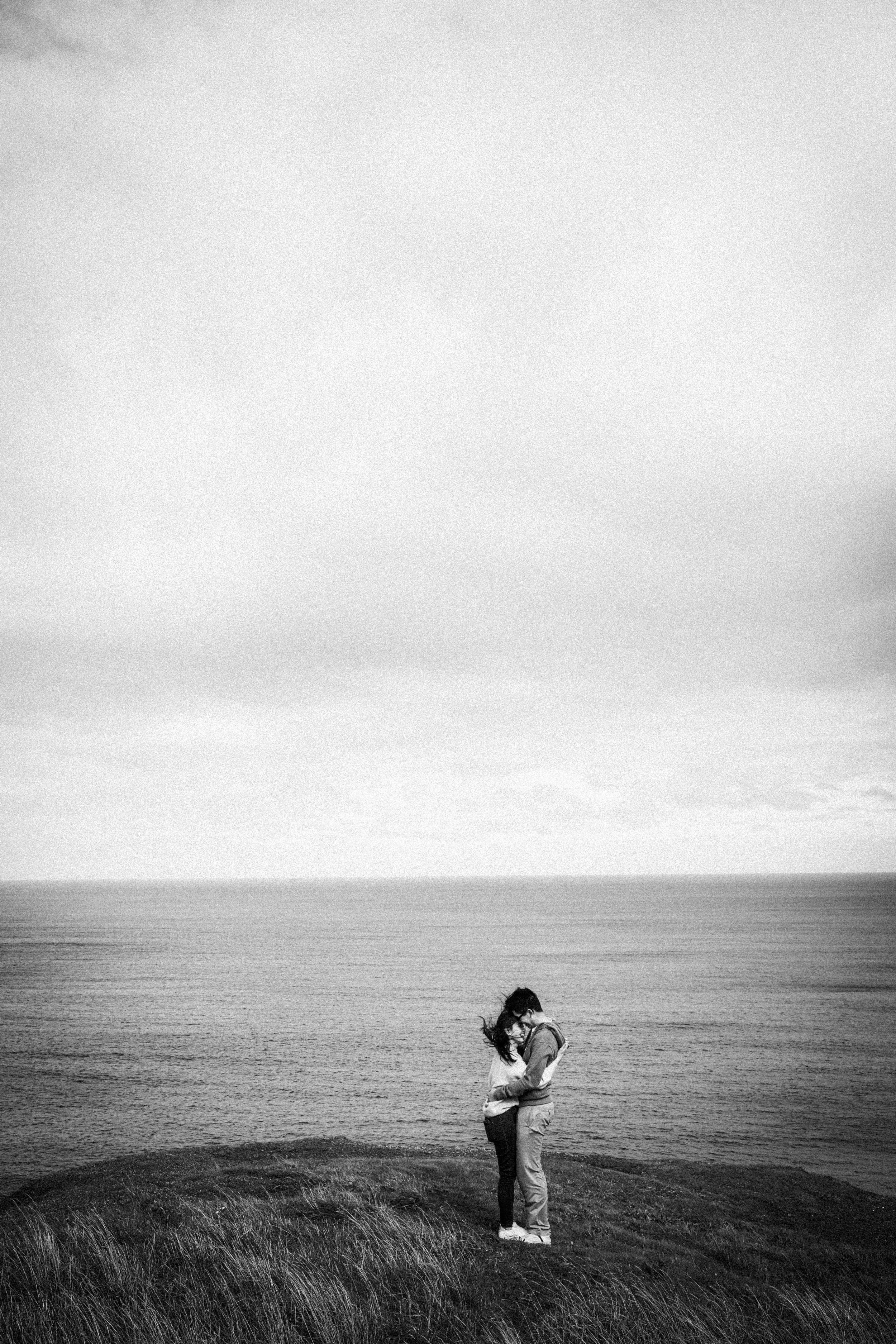 A couple hugging and kissing on a grassy area near the sea, with waves and a cloudy sky in the background in black and white.