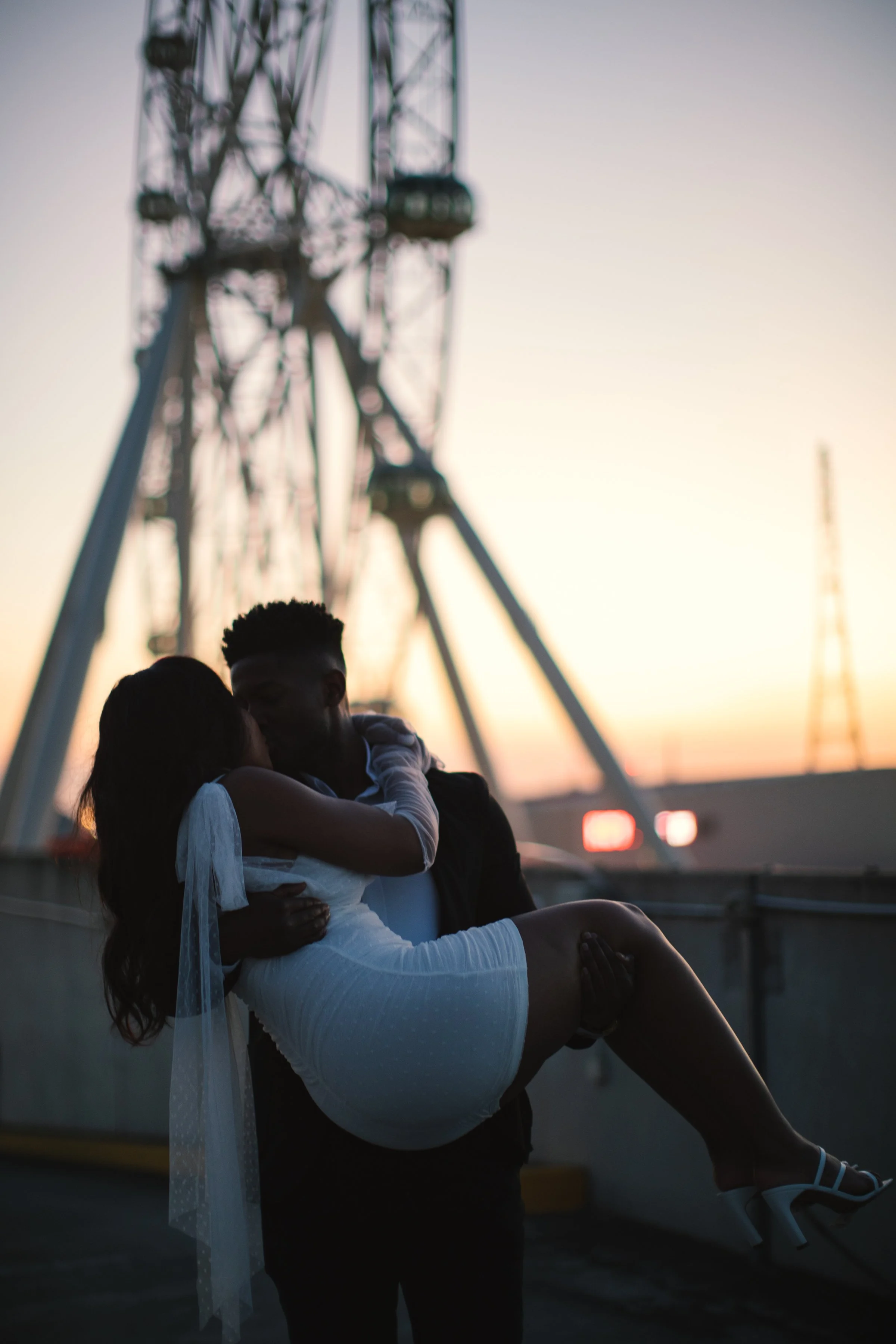A couple embracing each other on an amusement park ride during sunset, with a large Ferris wheel in the background.