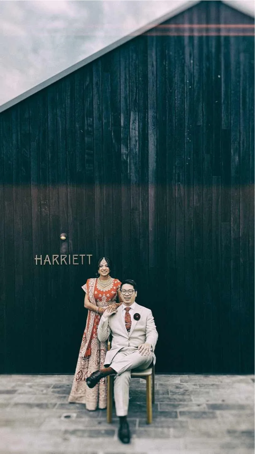 A woman in a traditional colorful dress and a man in a light-colored suit with a patterned shirt and tie, sitting on a chair in front of a dark wooden wall with the text "HARRIETT" on it. They are both smiling and holding hands.