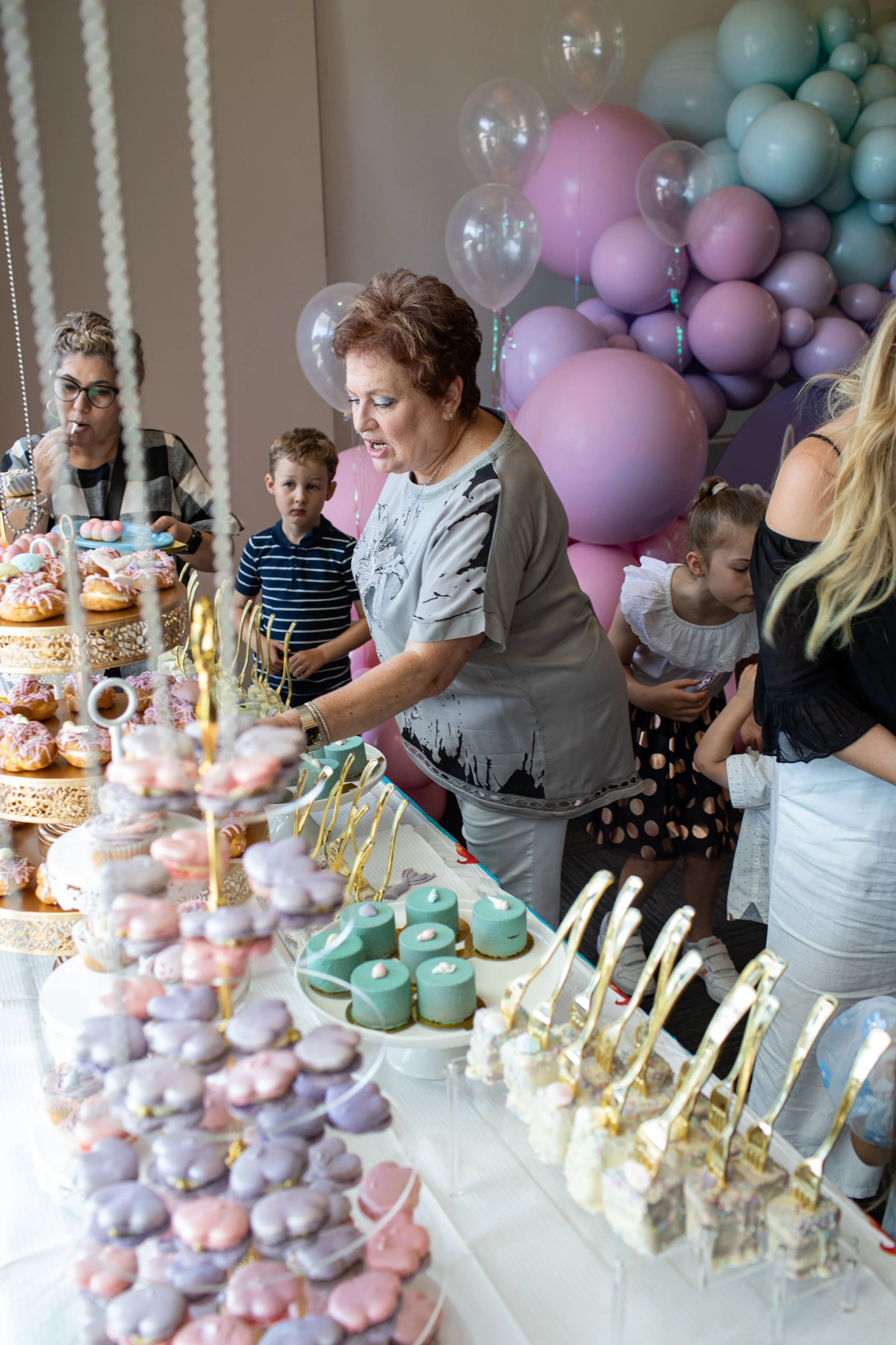 A dessert table filled with pink, purple, and green treats, including donuts, macarons, and popsicles, with people serving themselves at a celebration with pastel balloon decorations in the background.