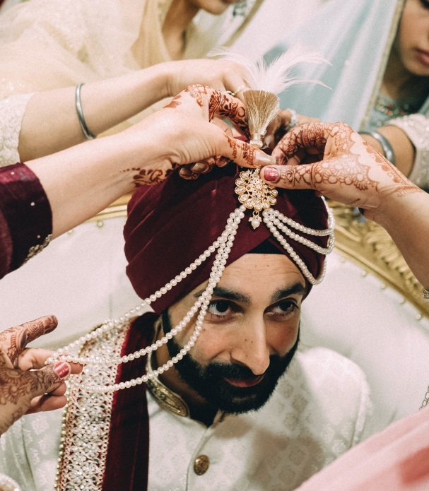 A groom in traditional Indian wedding attire having a turban ornament decorated with pearls and feathers placed on his head by women with hennaed hands.