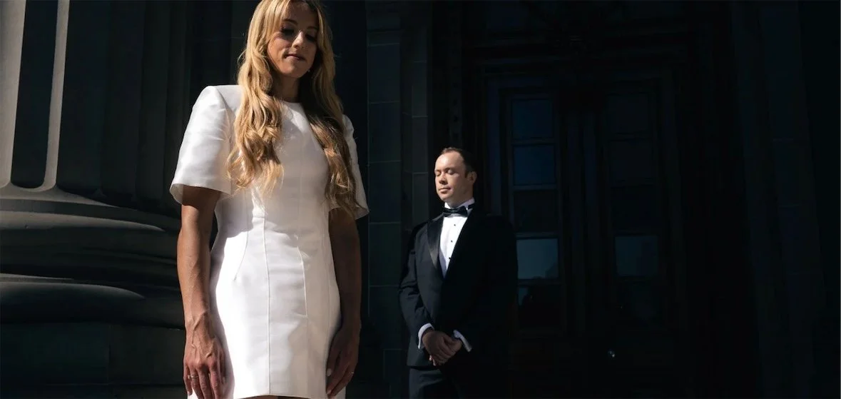 Woman in a white dress standing outdoors with a man in a black tuxedo in the background outside a dark building.