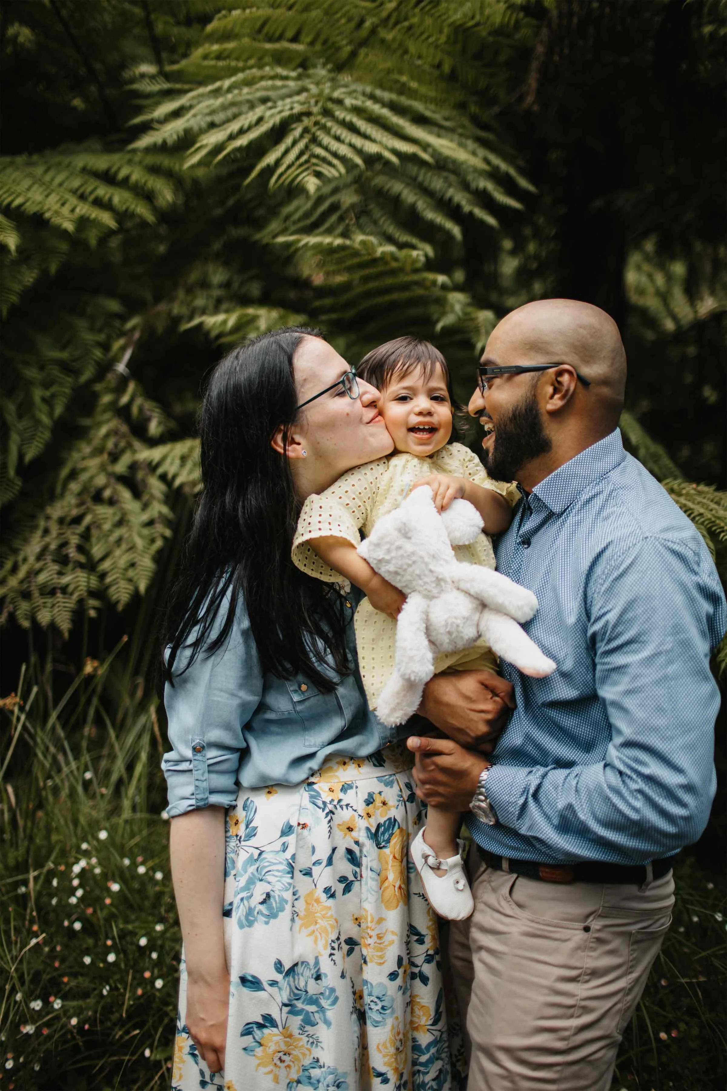 A family of three standing outdoors among lush green foliage, with the mother kissing the child on the cheek and the father smiling at them, holding the child in his arms.