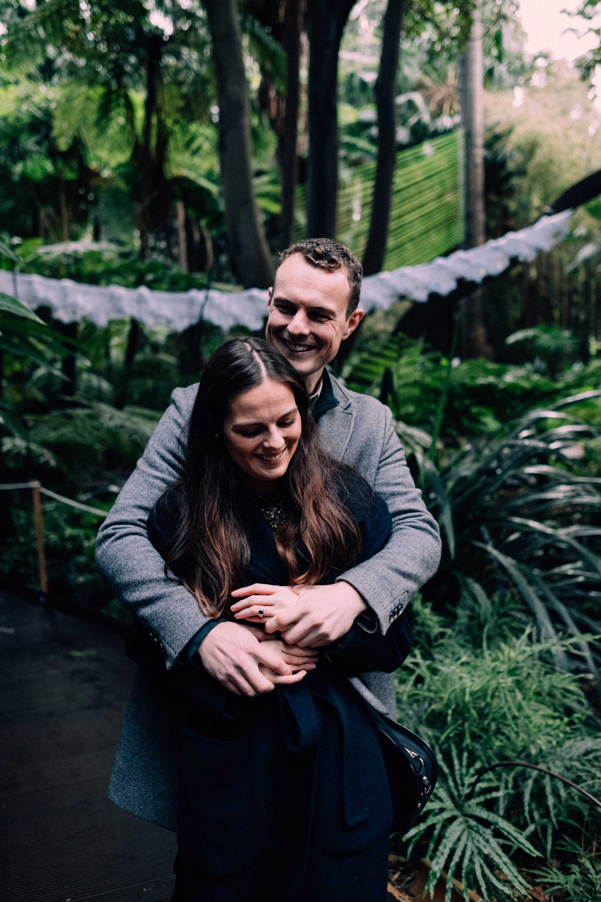 A man and a woman smiling and hugging in a lush, green indoor botanical garden with large plants and trees.