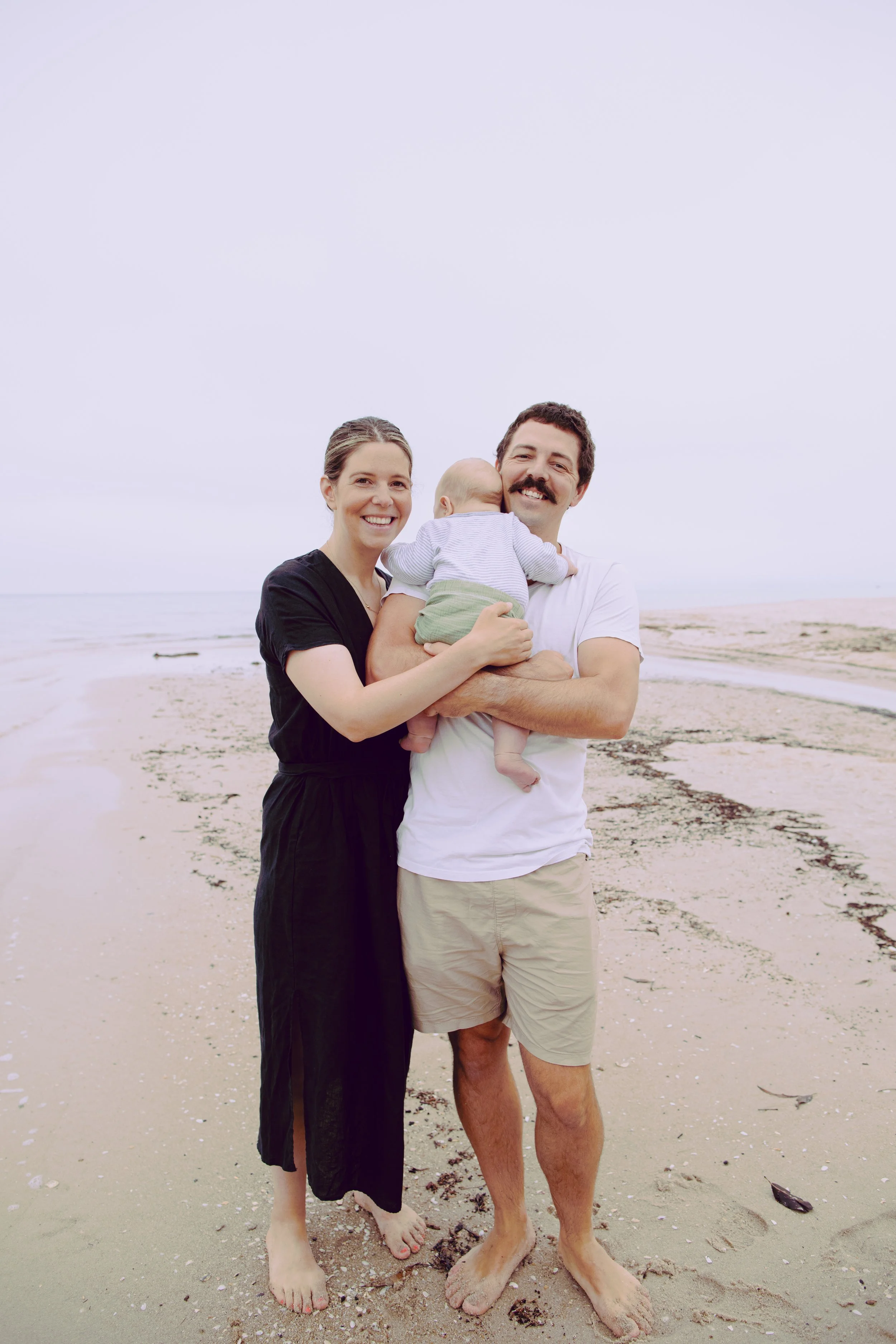 A family of three standing on the beach, smiling and embracing, with the ocean and sandy shore in the background.