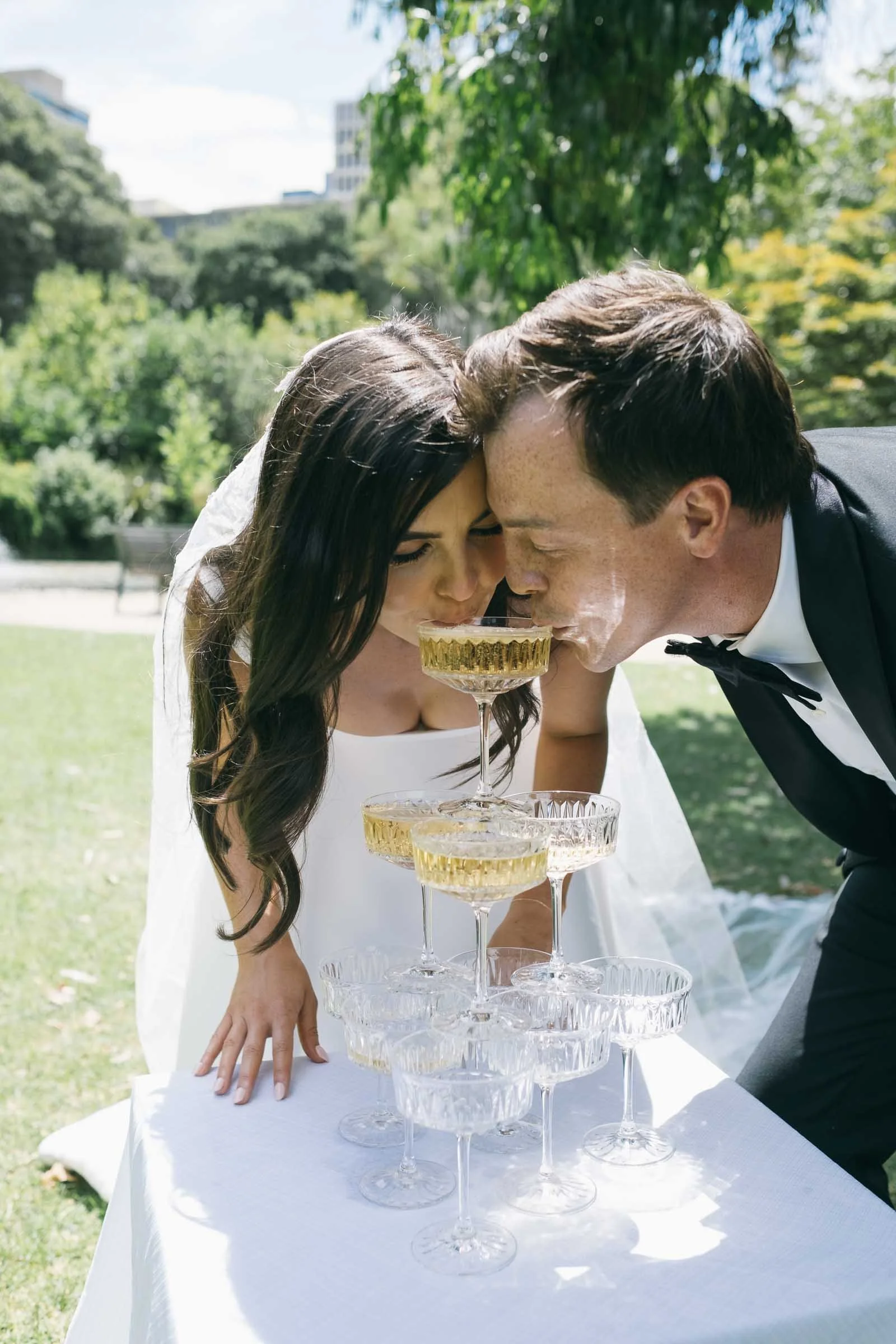 a couple having champagne from a small champagne tower in the treasury gardens in melbourne celebrating their elopement wedding.