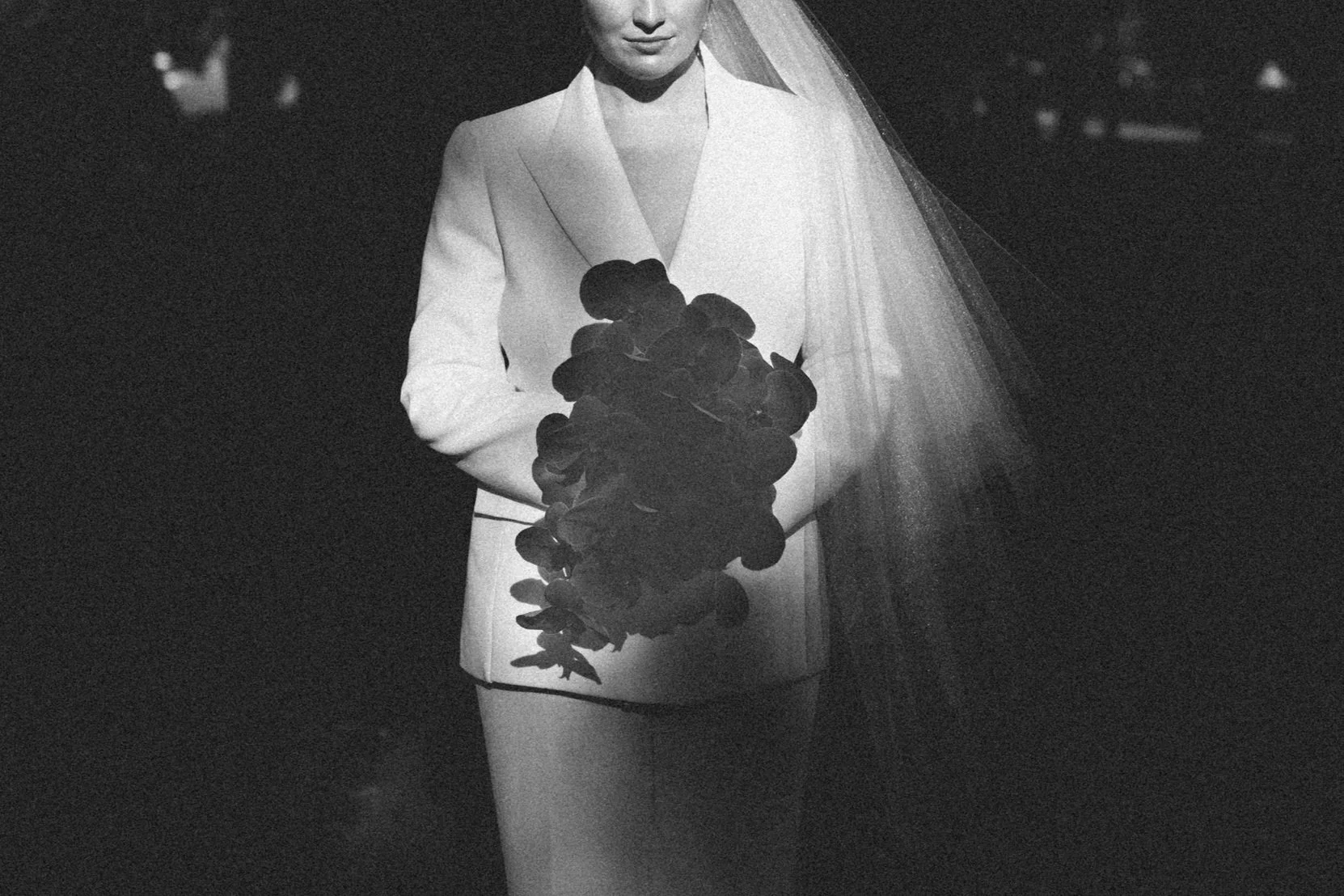 A woman in a white blazer and shorts is holding a bunch of dark flowers. She is wearing a veil, and the background is dark.