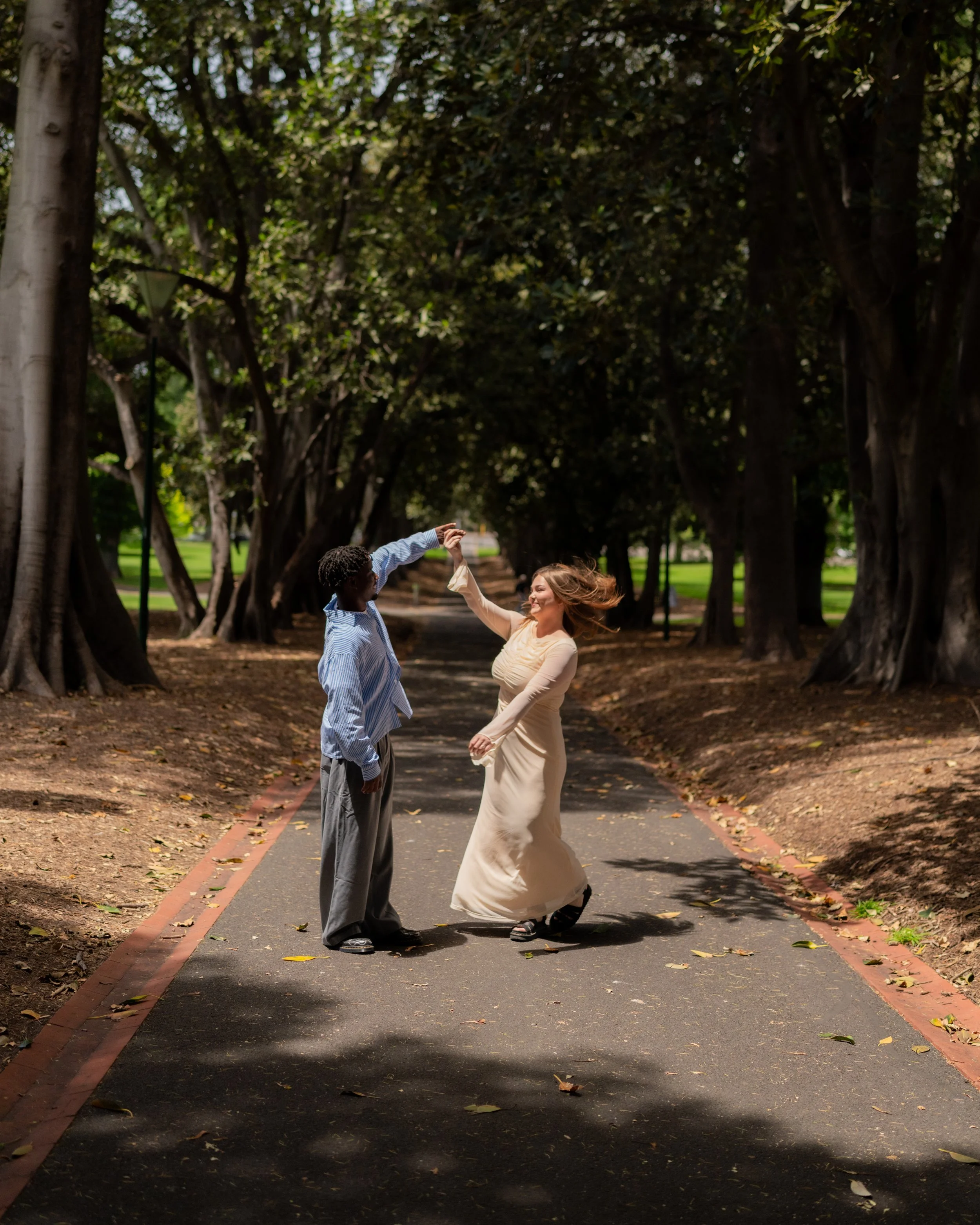 A man and woman dancing together in a park with trees and a walking path, holding hands and spinning around.