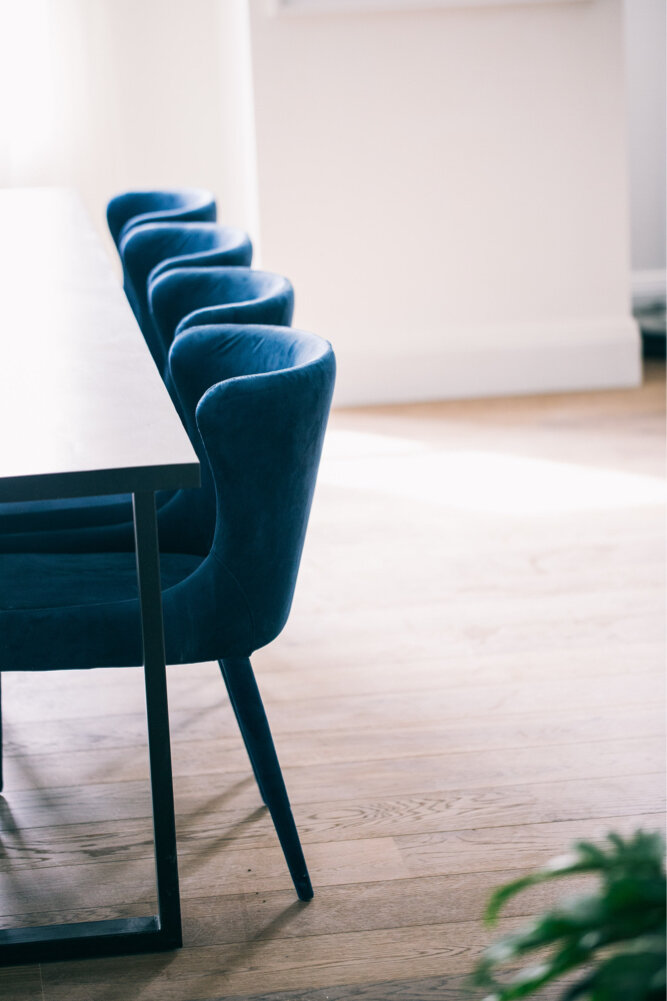 Blue upholstered dining chairs lined up next to a wooden table in a sunlit room with wooden flooring.