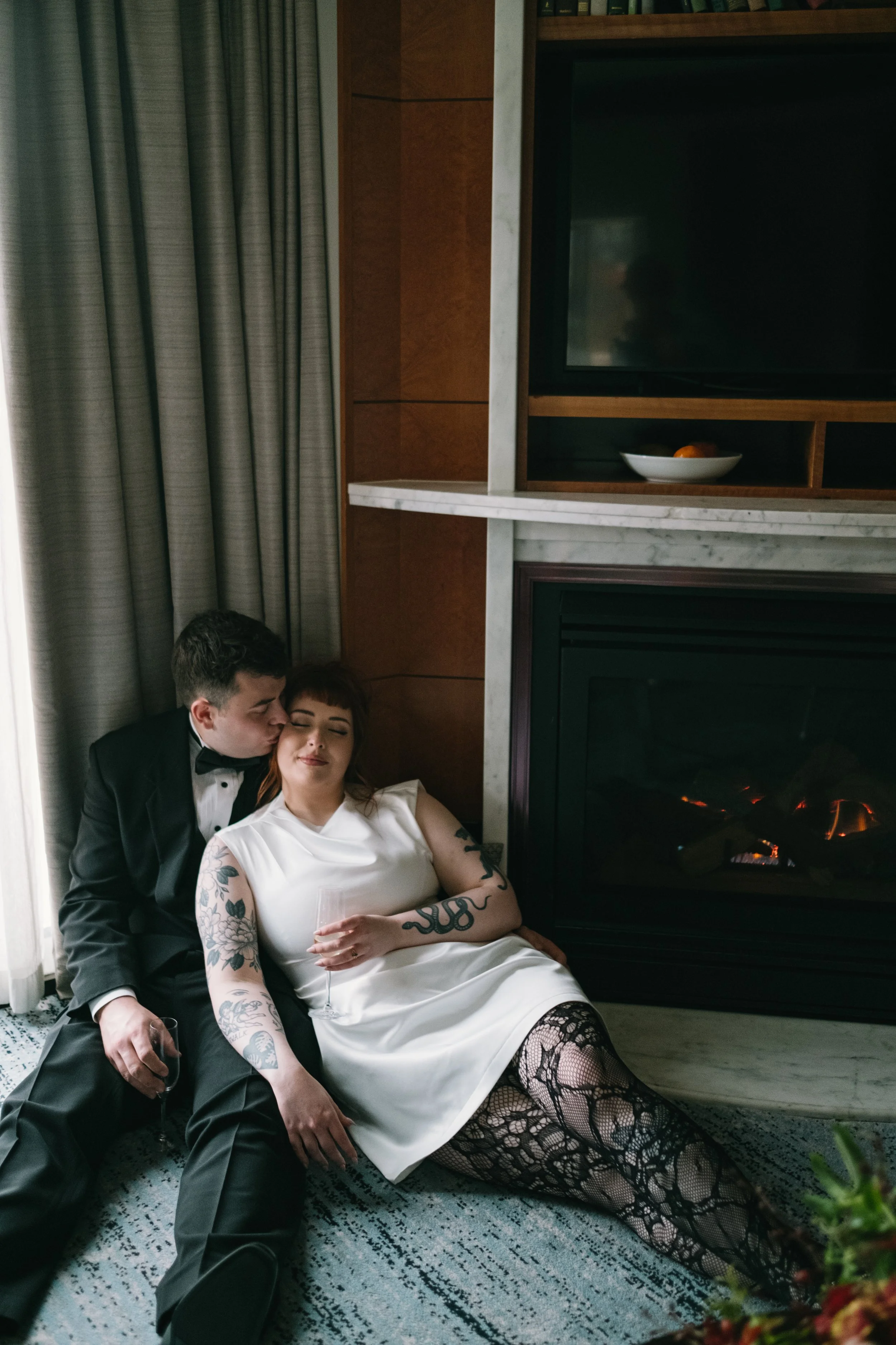 A couple dressed in formal attire relaxing by a fireplace. The man is in a tuxedo, and the woman is in a white dress with lace stockings, holding a glass of wine.